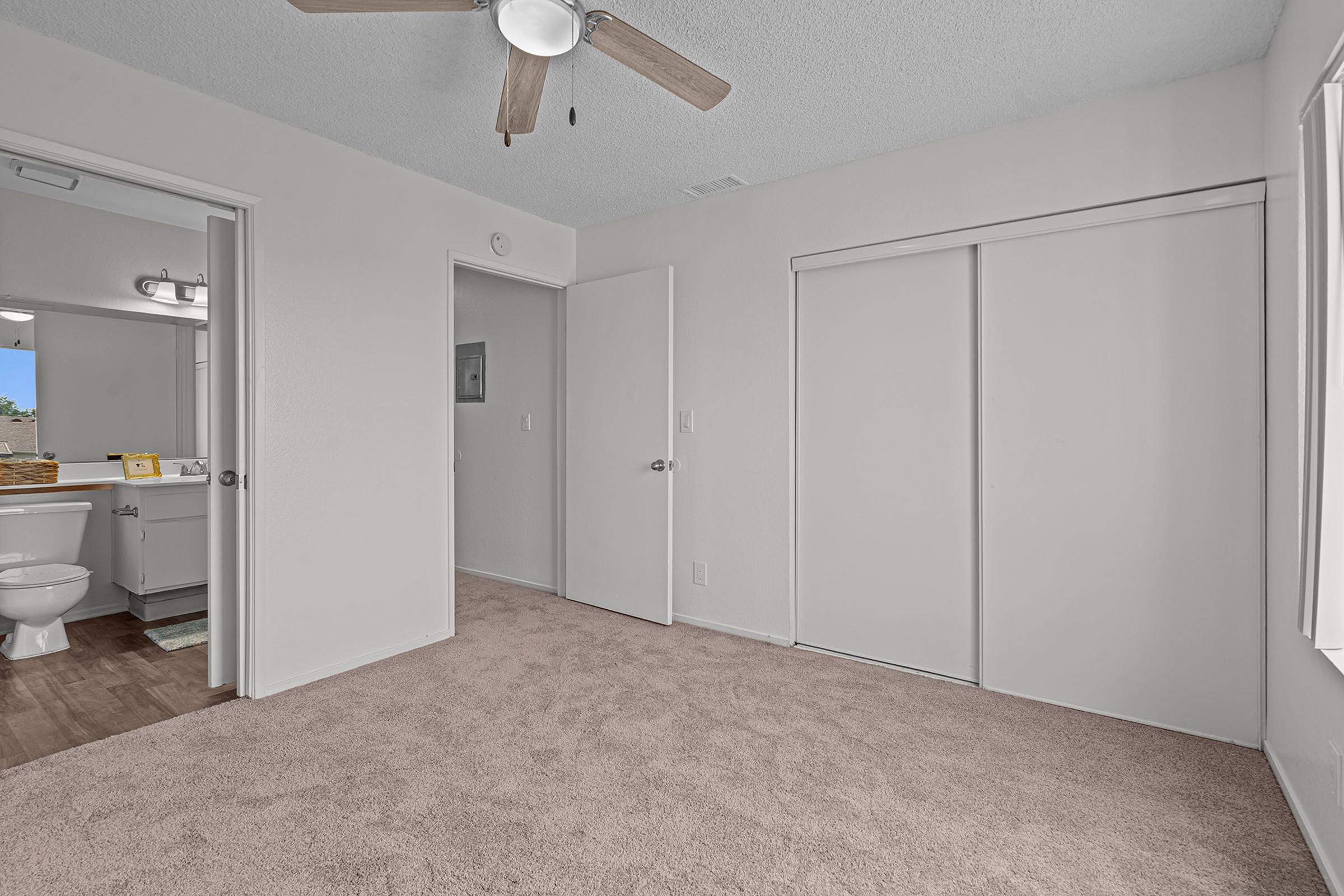 A clean, well-lit bedroom featuring light-colored walls and plush beige carpet. The room has a ceiling fan, a closet with sliding doors, and an open doorway leading to a bathroom. Sunlight enters through a window, creating a bright atmosphere.