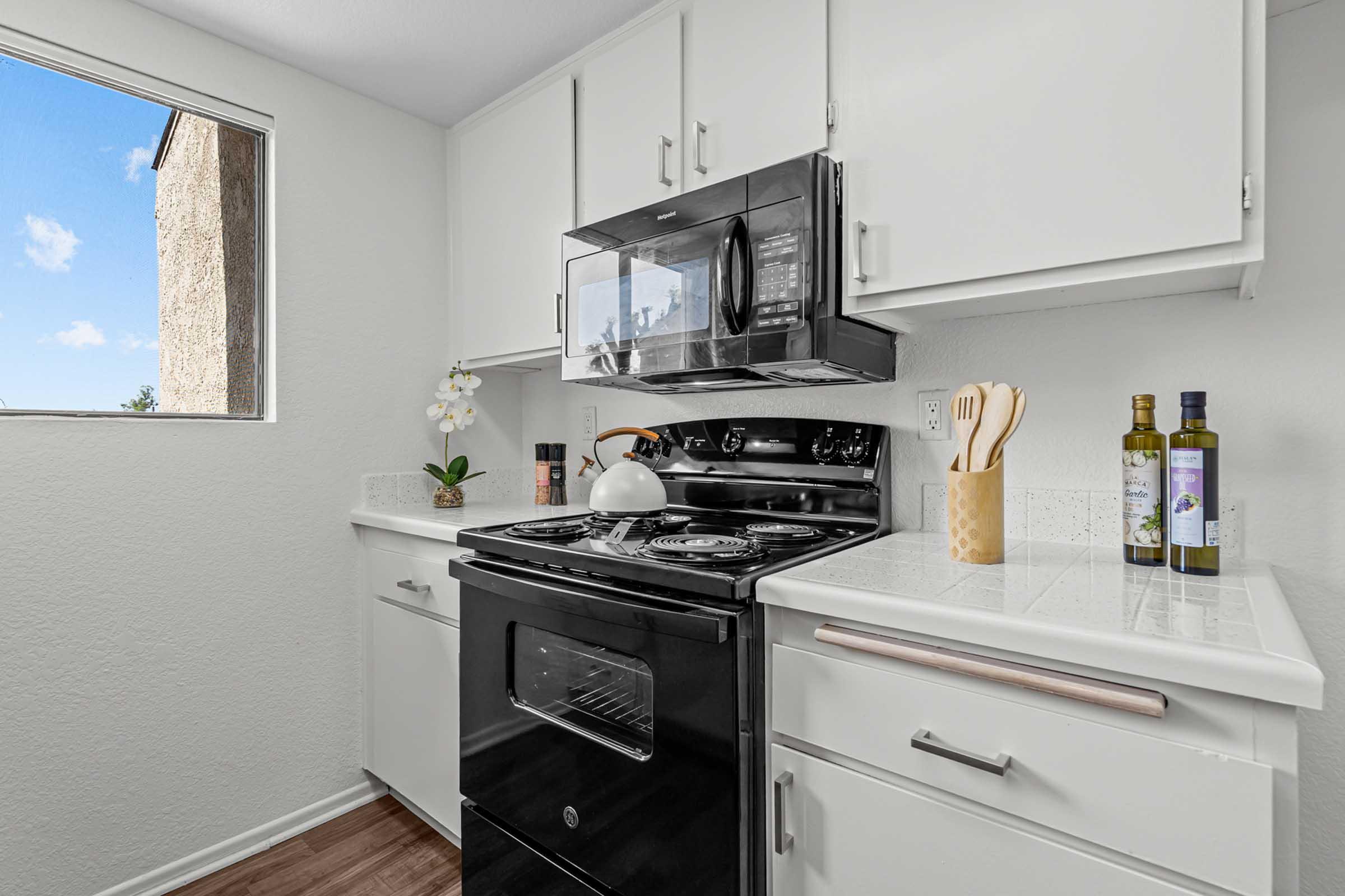 Modern kitchen featuring a black stove and microwave, white cabinetry, and a clean countertop. There are cooking utensils in a holder, olive oil bottles, and a decorative plant. A window allows natural light to brighten the space, highlighting the minimalist design and contemporary feel.