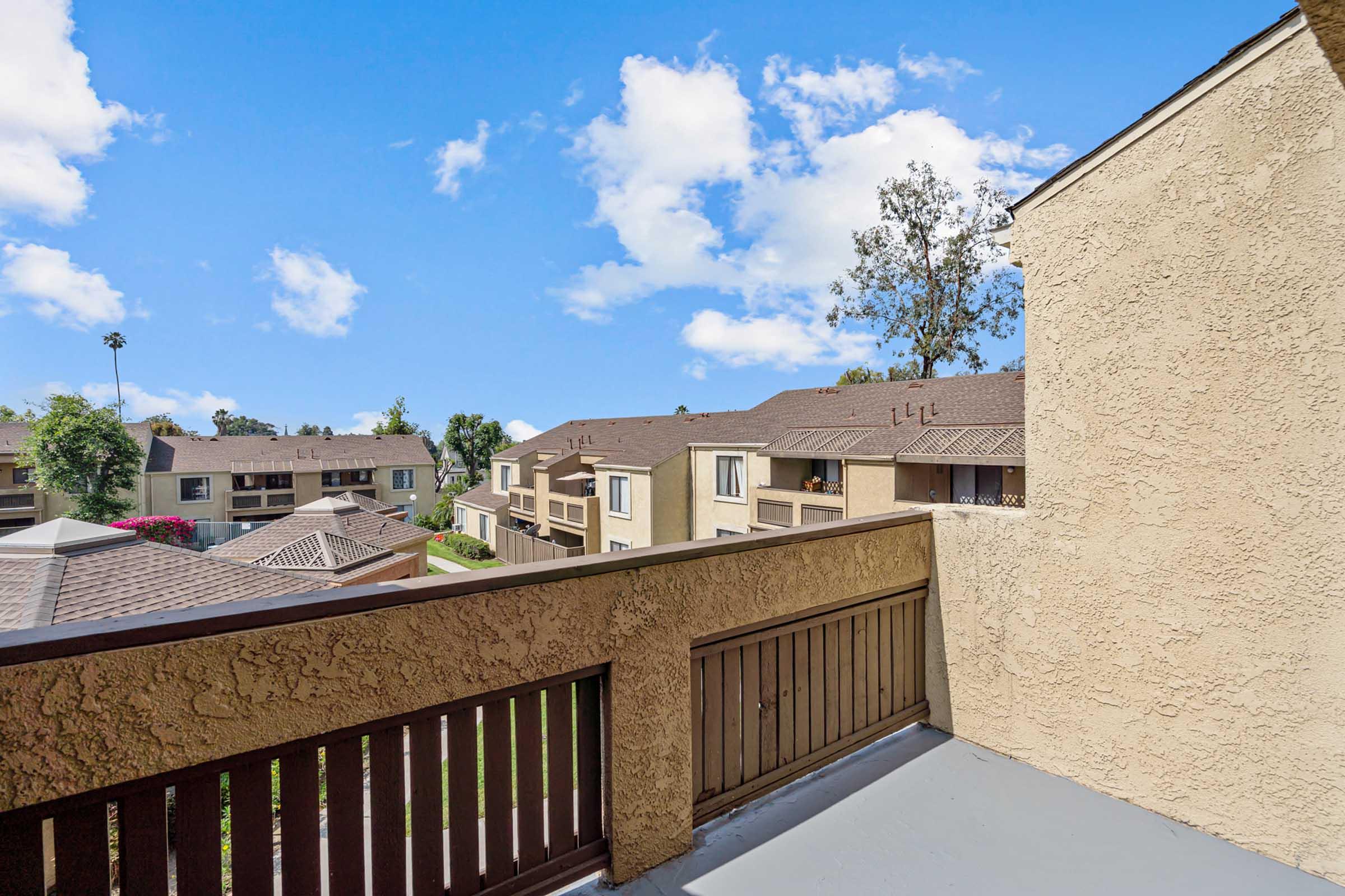 View from a balcony overlooking a residential area, featuring several multi-story apartment buildings, green trees, and blue sky with fluffy white clouds. The scene conveys a serene suburban atmosphere.