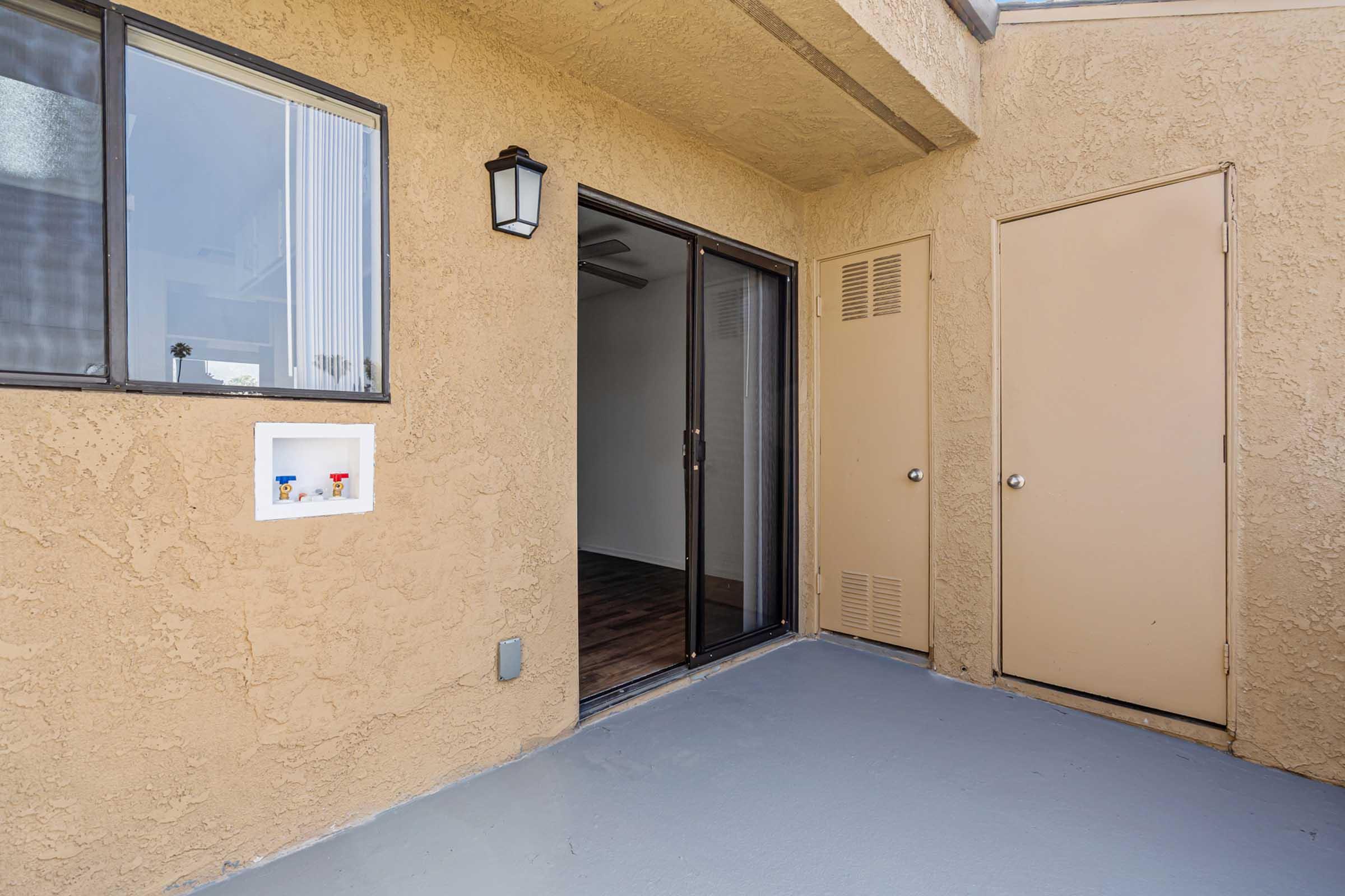 A view of an entrance to an apartment featuring a sliding glass door, beige walls, and a small window. There is a wall-mounted lantern beside the door, and a closed door on the right. The floor is painted gray, and the interior appears empty.