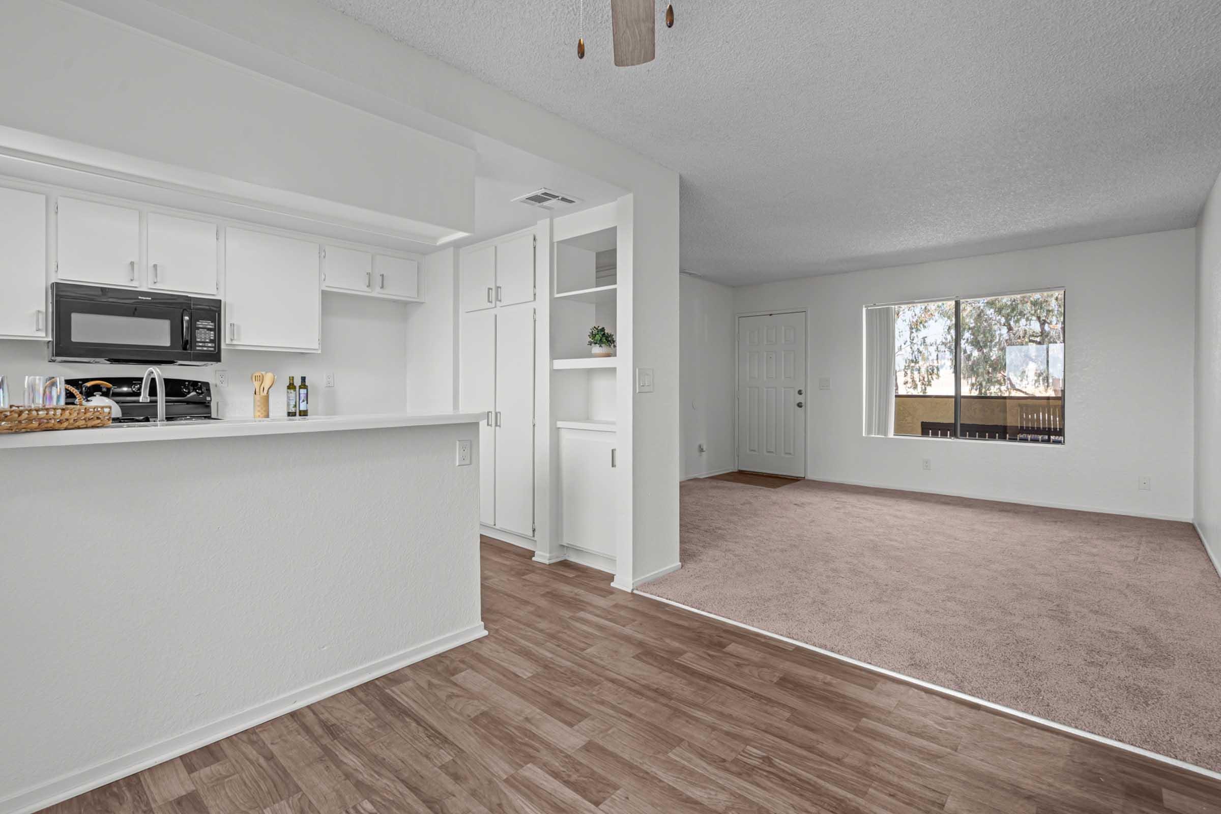A bright, modern living space featuring light-colored walls, a kitchen area with white cabinetry and a black microwave, and a spacious carpeted living room. A large window lets in natural light, showcasing a view of trees outside. The layout conveys an open and inviting atmosphere.