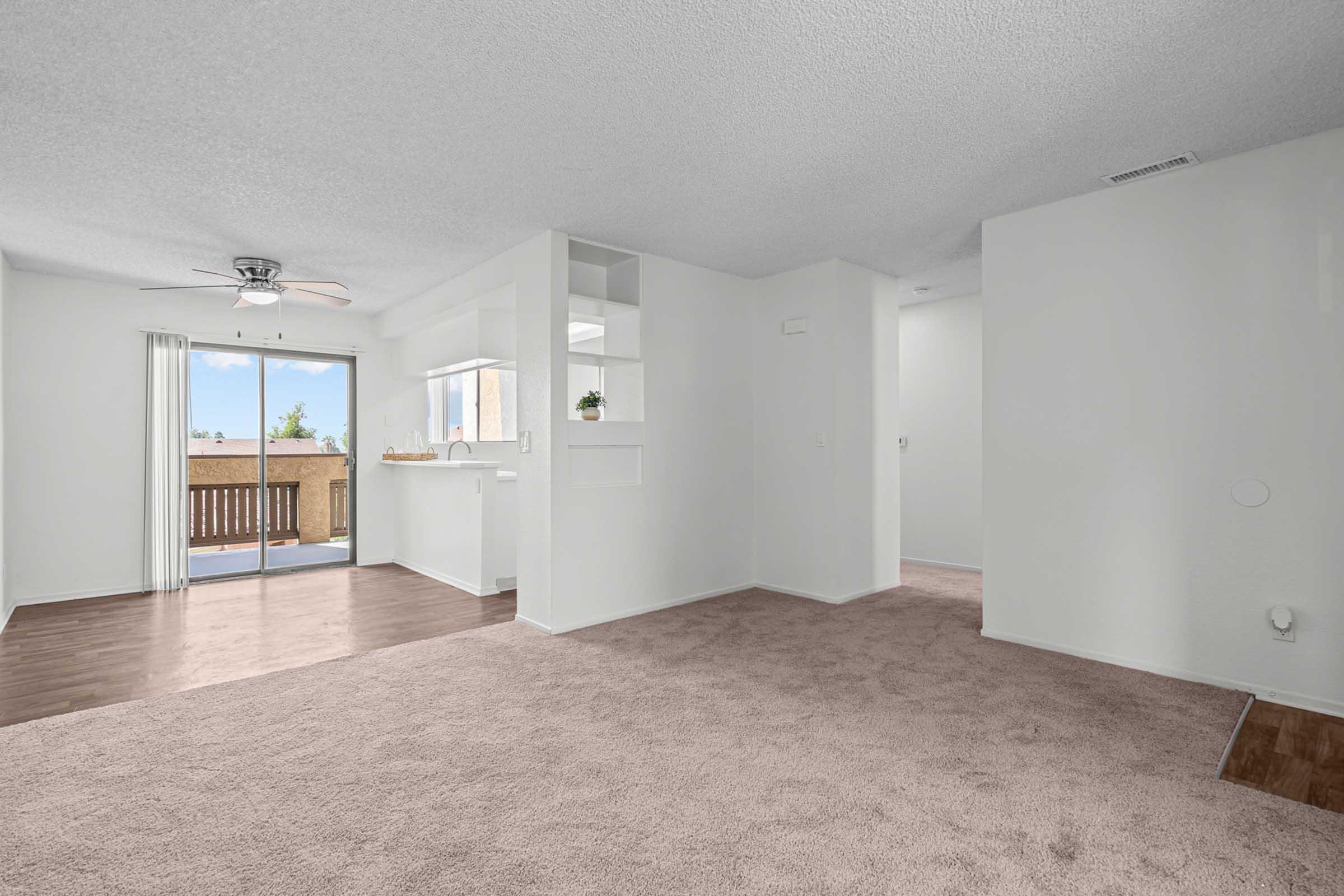 Bright and spacious living area with light-colored walls, plush carpet, and large windows allowing natural light. Includes a ceiling fan and a sliding glass door leading to a balcony. The adjacent area features a small open kitchen with modern fixtures. The overall ambiance is inviting and well-lit.
