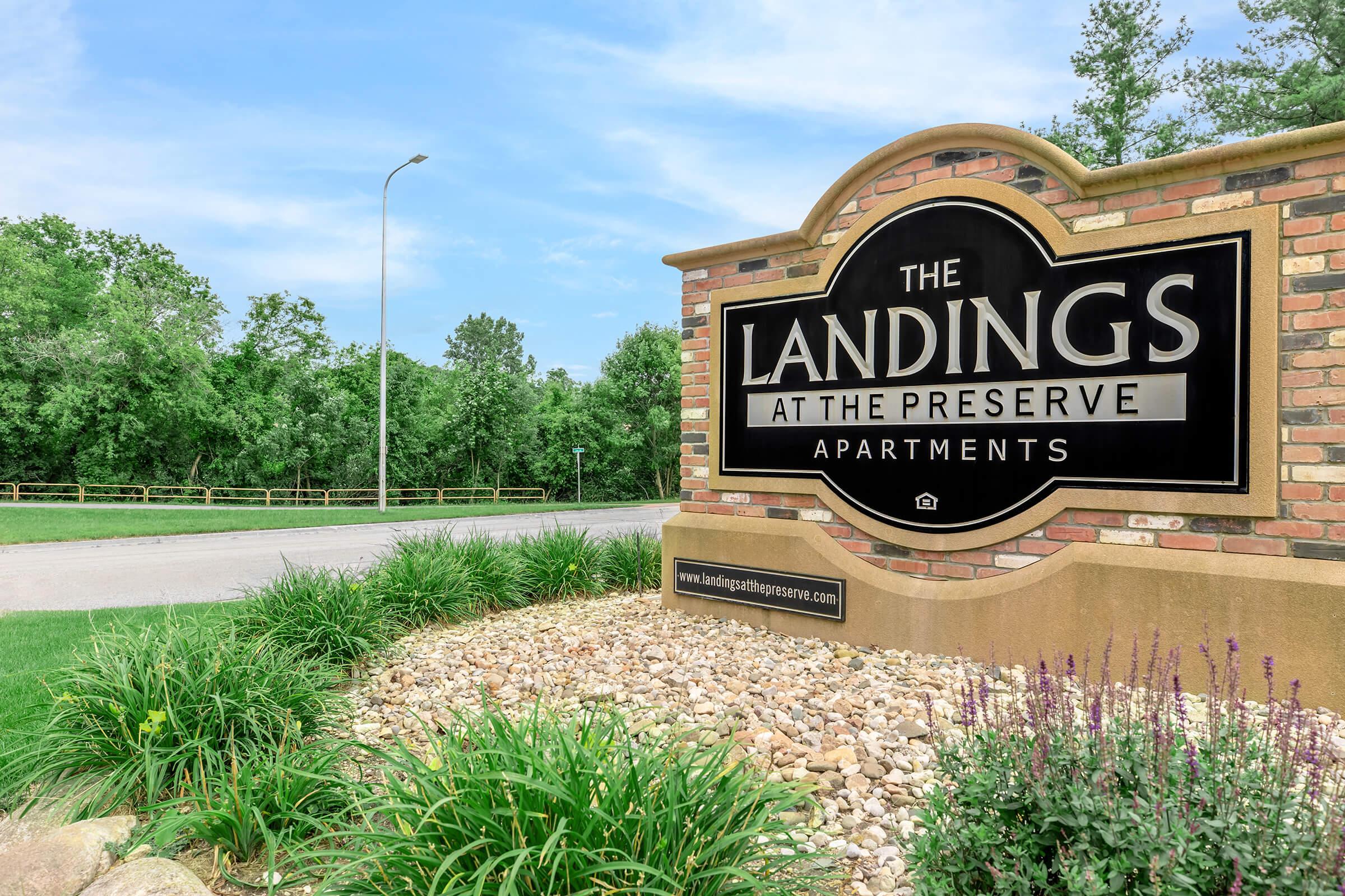 Sign for "The Landings at the Preserve Apartments" with lush greenery and a gravel landscape in the background.