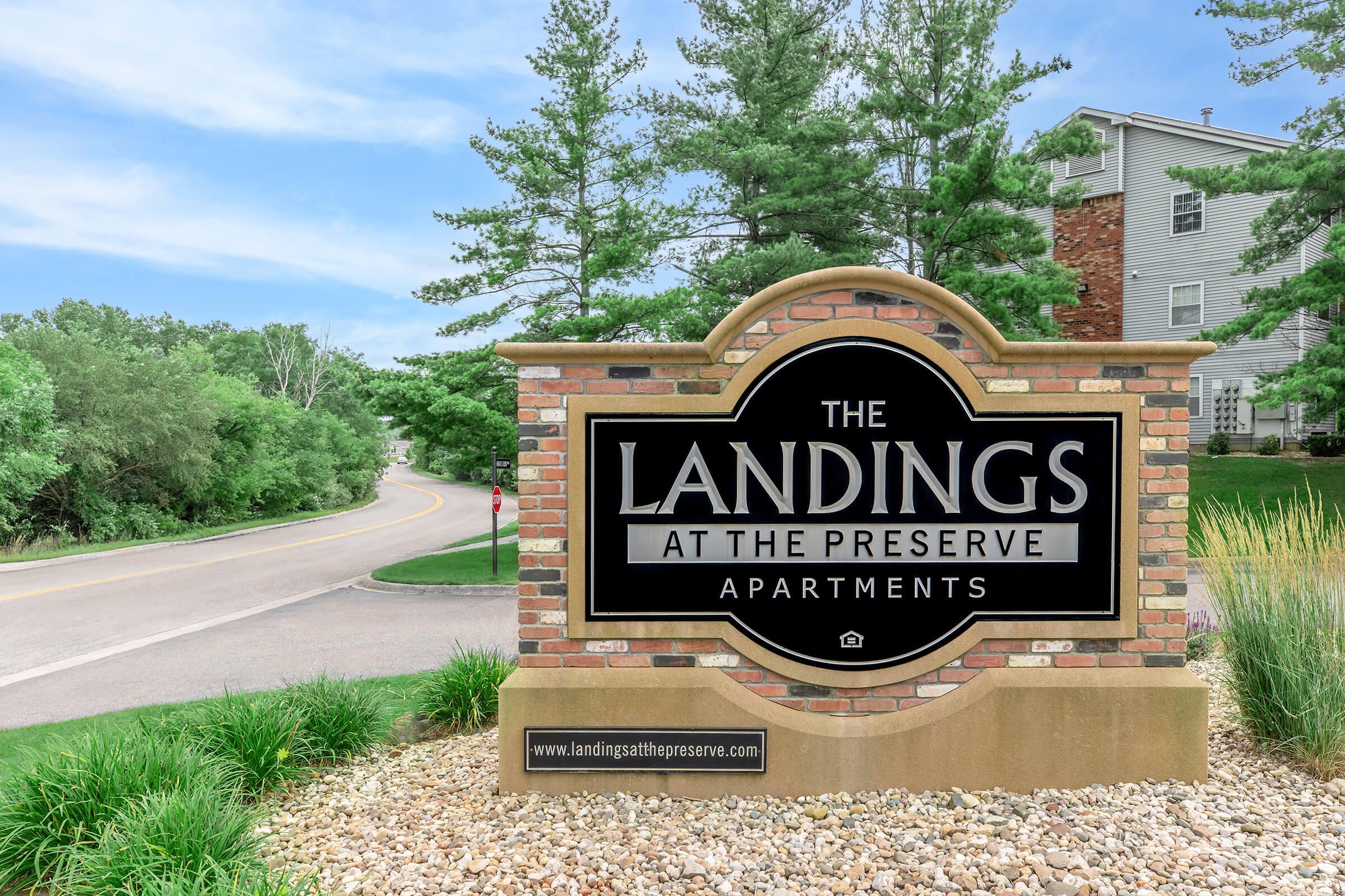 Sign for "The Landings at the Preserve Apartments" featuring a brick design, set among green trees and a roadway in the background.