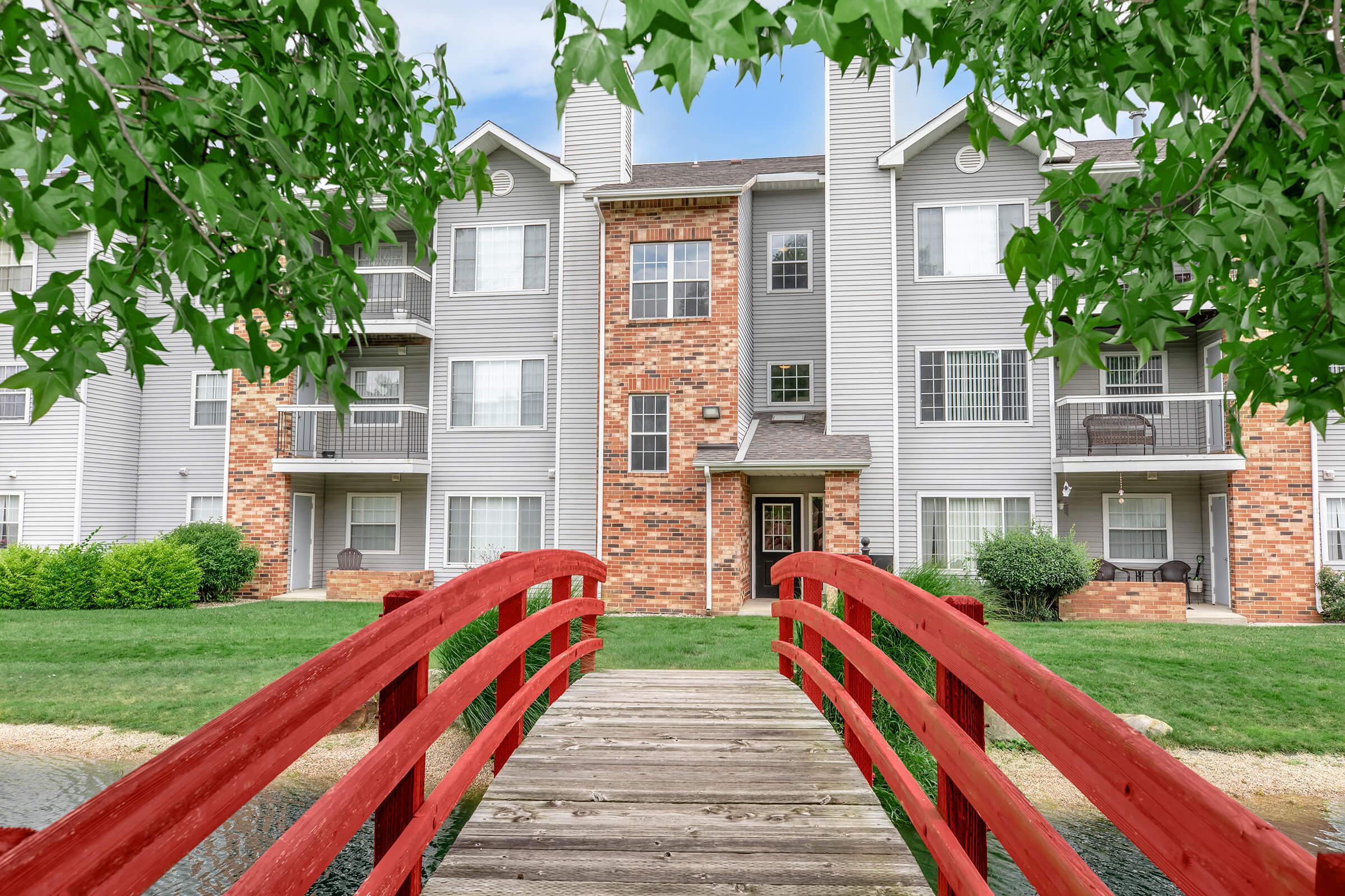 A red wooden bridge leading to a multi-story residential building with a combination of brick and gray siding, surrounded by green grass and shrubs.