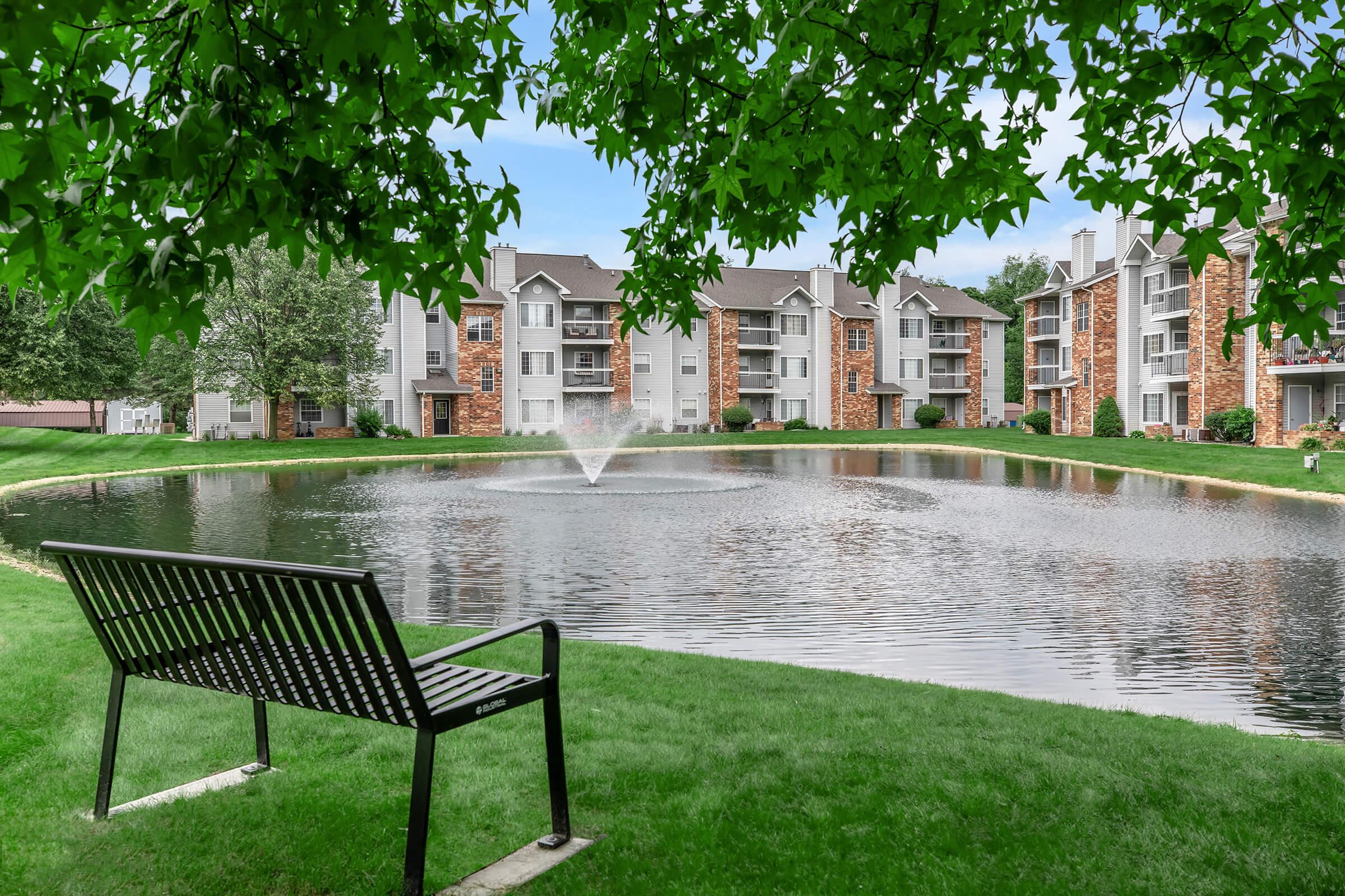 A serene pond surrounded by lush green grass and a bench in the foreground, with apartment buildings visible in the background and a fountain gently spraying water in the center of the pond.