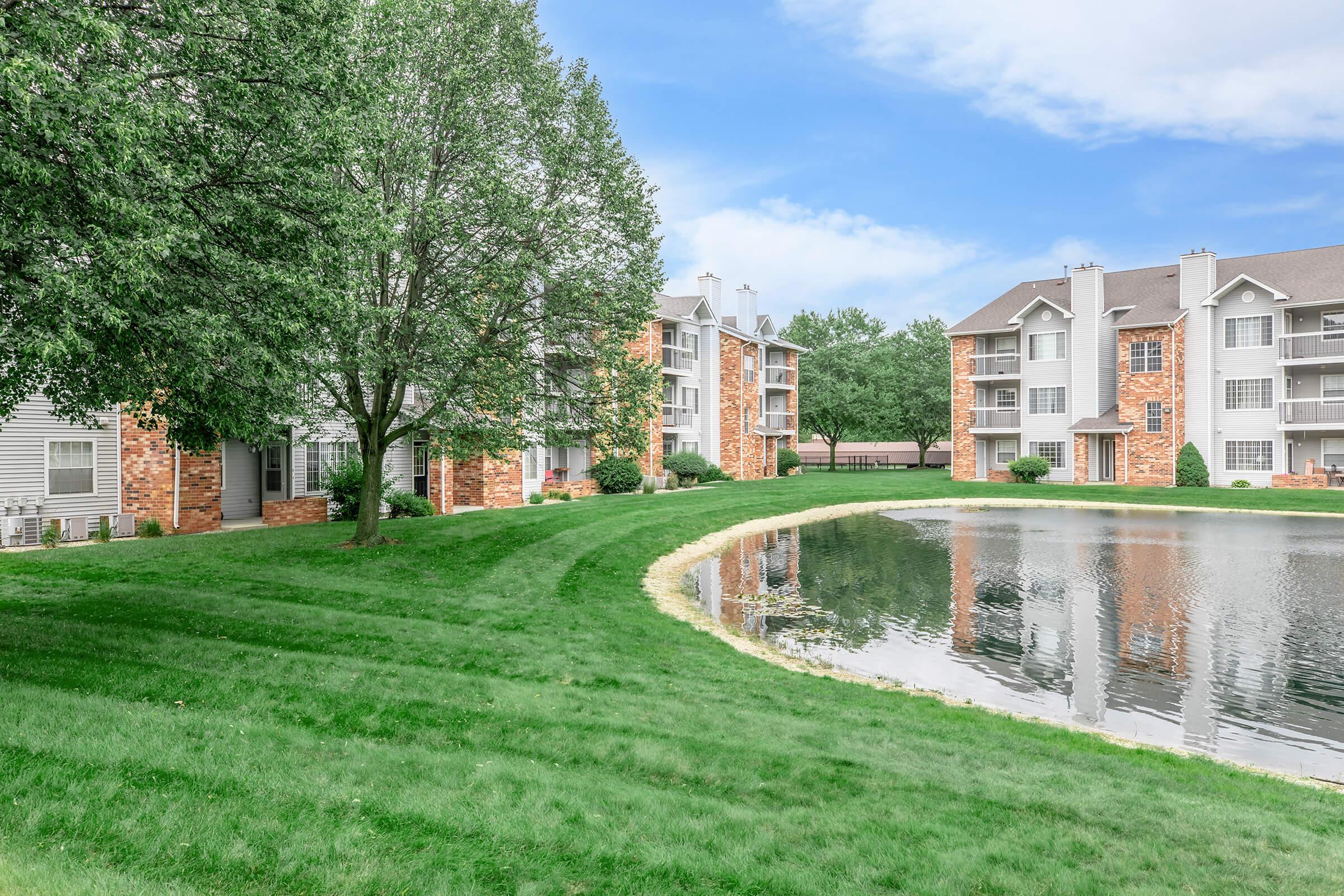 A serene landscape featuring a well-maintained lawn, a tranquil pond, and apartment buildings in a residential area. The sky is partly cloudy, and trees provide shade around the pond.