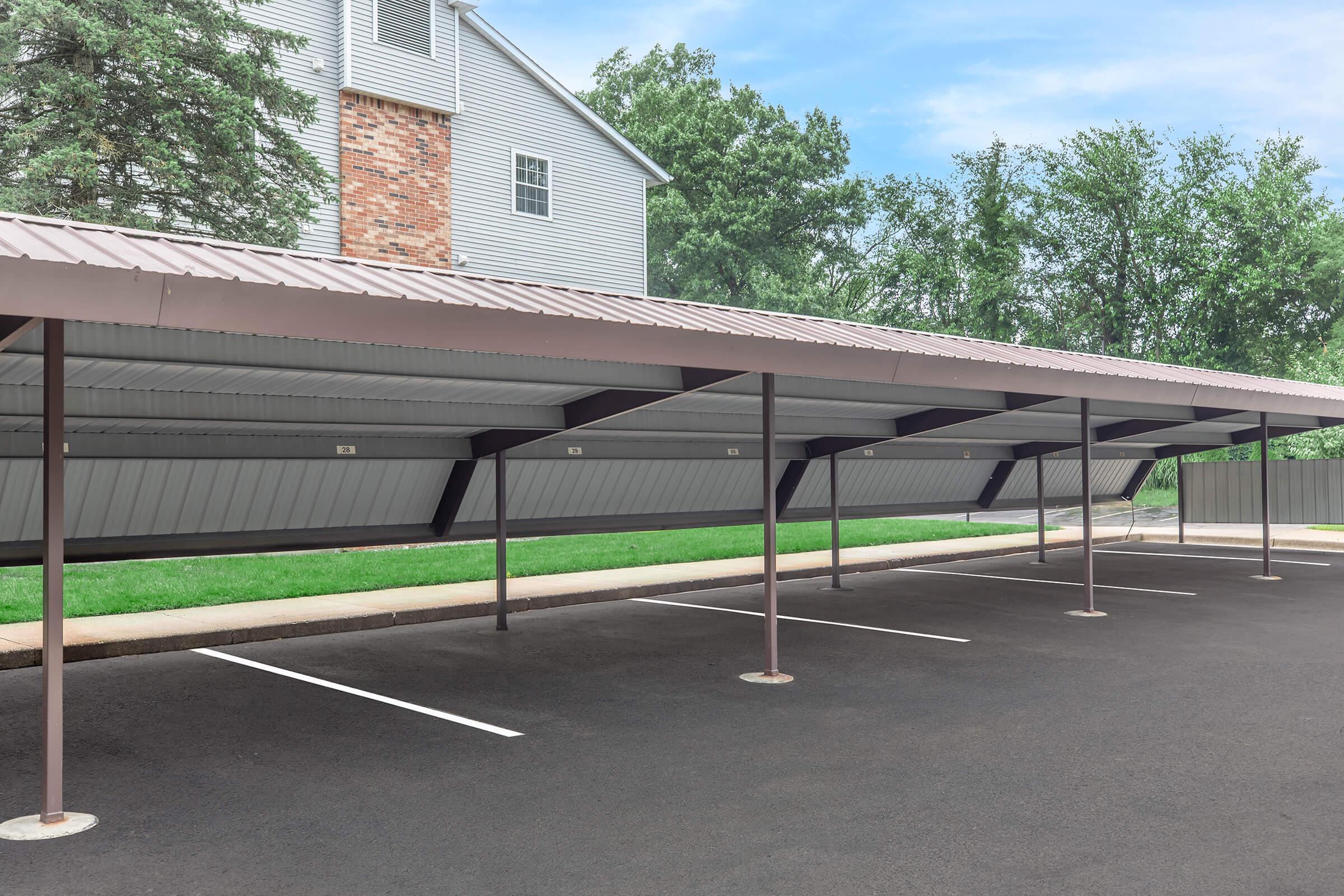 A covered parking area with a metal roof, several empty parking spaces, and grassy landscaping in the background. The structure has a modern design with a brick building partially visible in the rear.