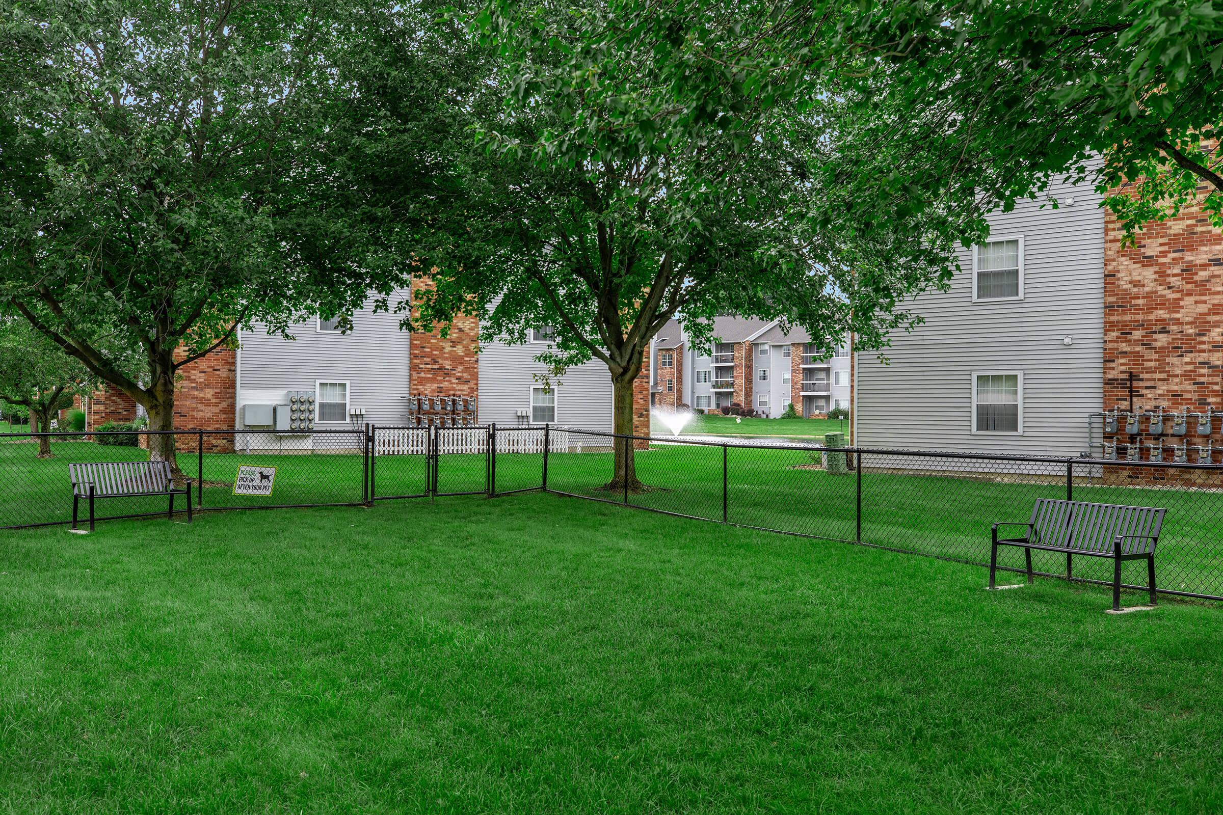 A green grassy area with a fenced-in space, featuring benches and trees, surrounded by residential apartment buildings in the background.