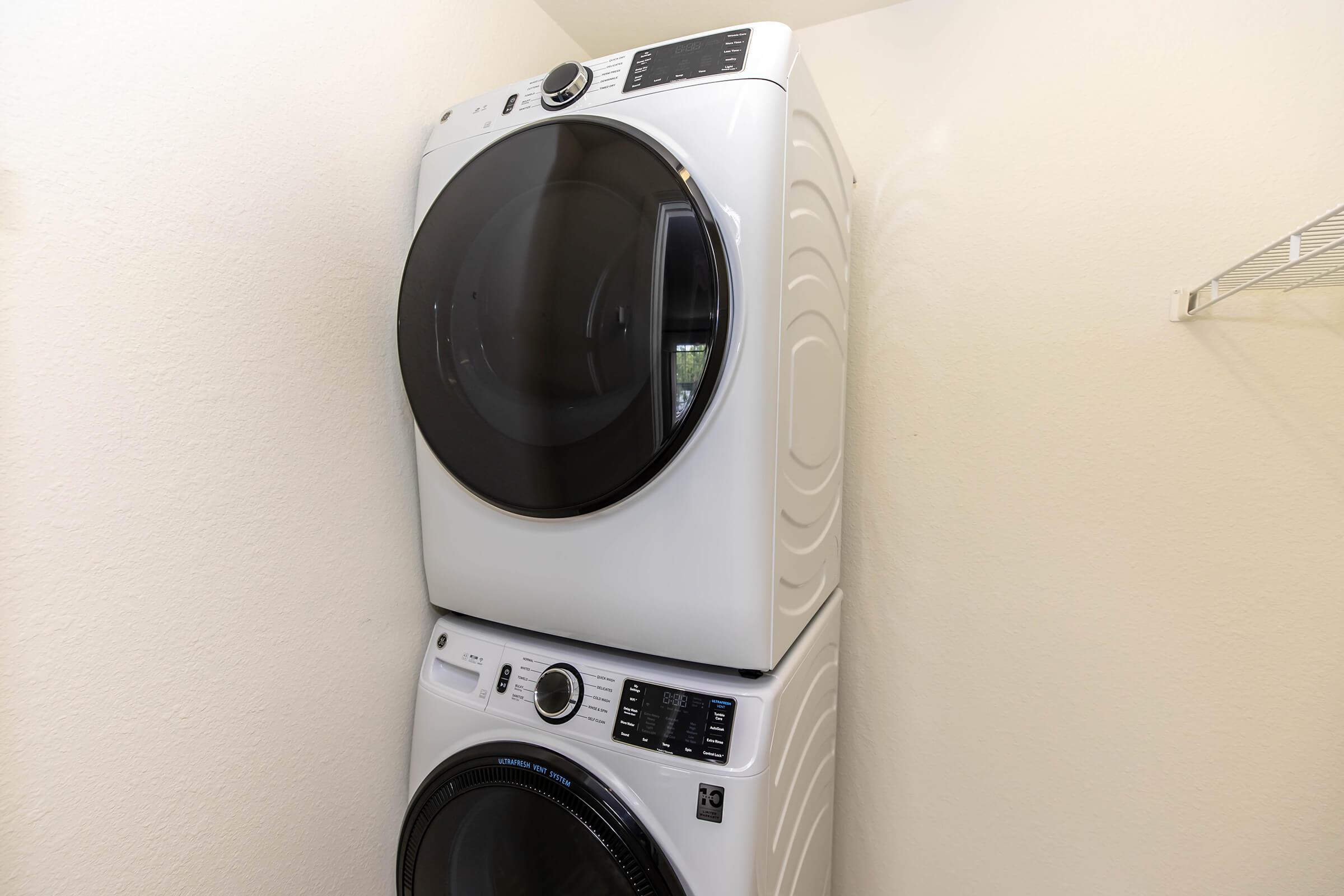 A white stacked washer and dryer unit in a corner of a laundry space. The washer is on the bottom, and the dryer is above. The machines have modern controls and are placed against a light-colored wall, with a shelf above for storage.