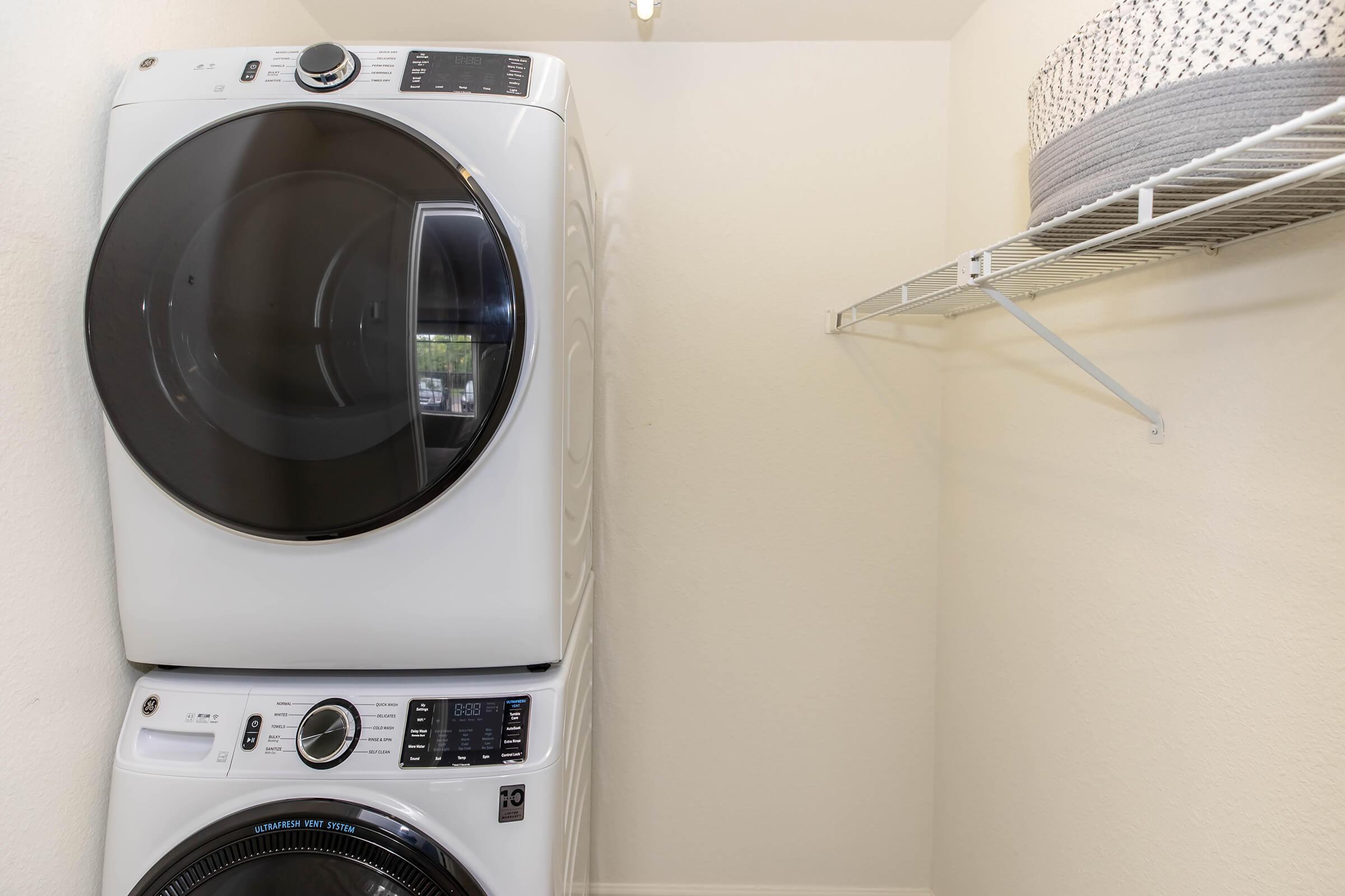 Stacked washer and dryer units located in a laundry nook, with a wire shelf above. The walls are painted a light color, and the space is well-lit, creating a clean and functional laundry area.