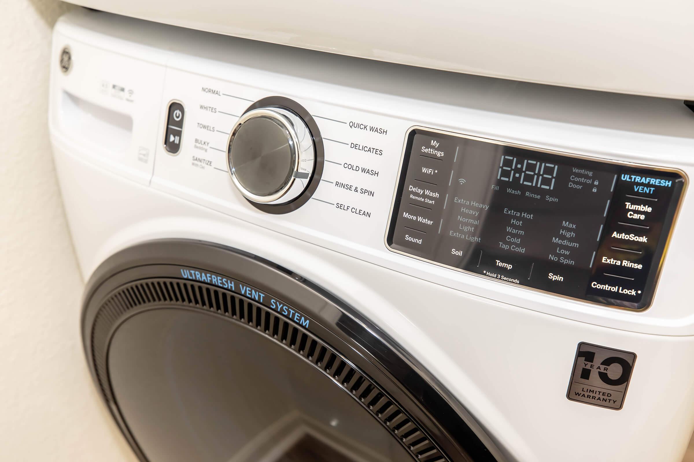 A close-up view of a modern washing machine's control panel displaying various settings and options, including a dial for selecting wash cycles, digital display for time, and buttons for features like "Tumble Care" and "Auto Soak." The machine has a sleek design with a large circular door.