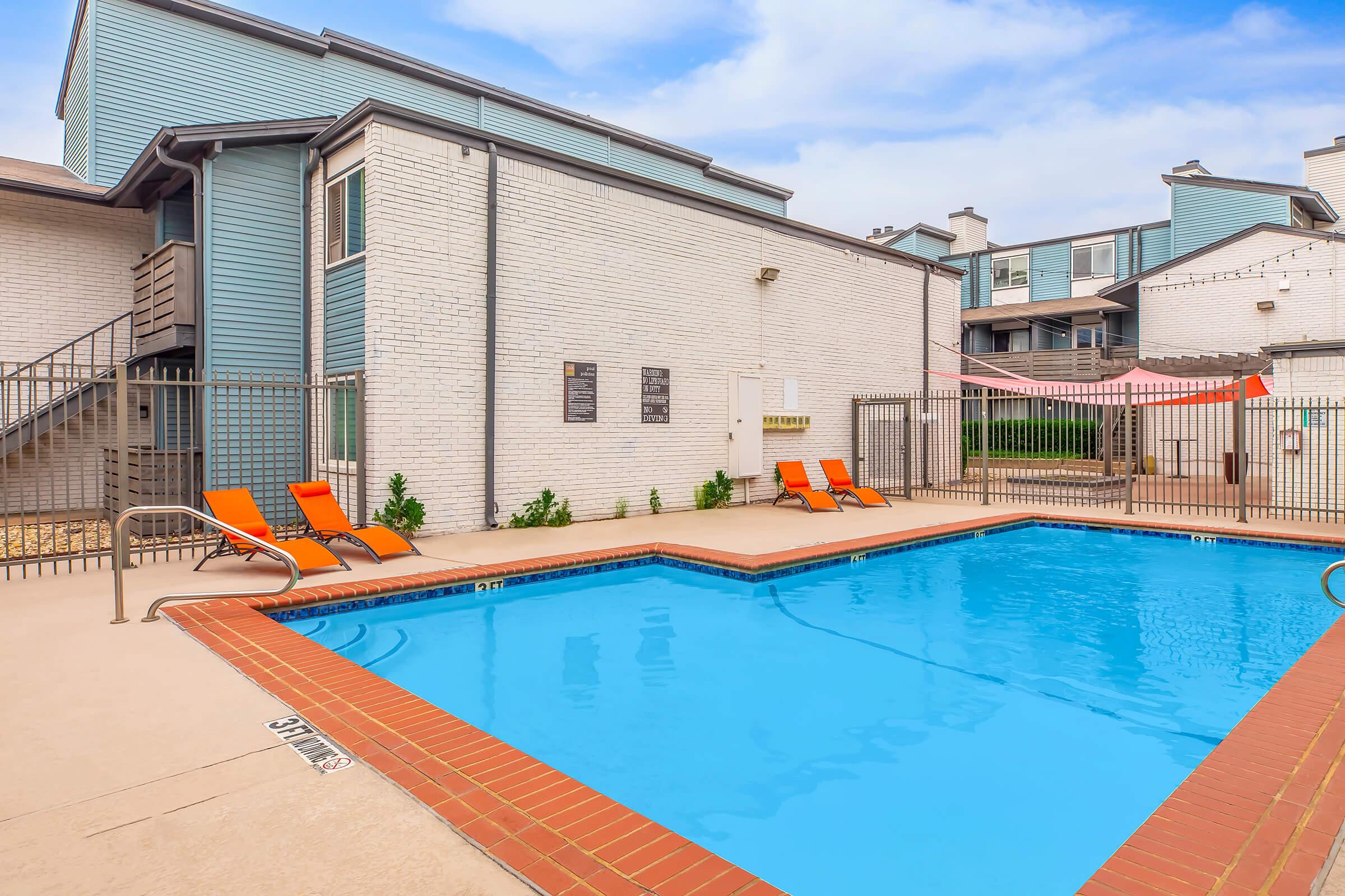 A clear swimming pool surrounded by a tan deck, with three orange lounge chairs positioned nearby. In the background, a light-colored building features a mix of textures and colors, and a fence encloses the pool area. The sky is partly cloudy, contributing to a bright, inviting atmosphere.