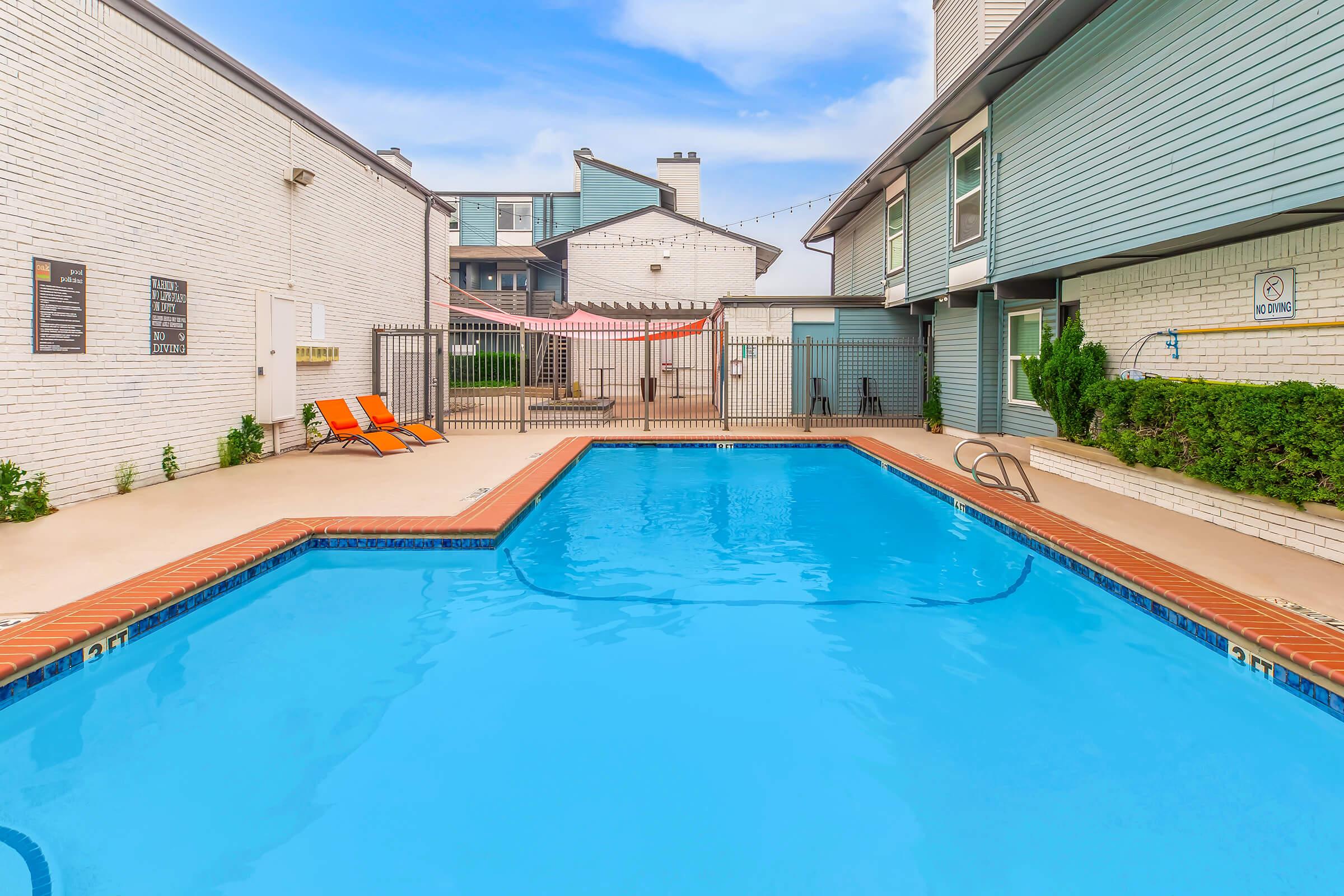A clear blue swimming pool surrounded by a brick deck, with two orange lounge chairs on one side. Enclosed by a black fence, the pool area features a well-maintained, light-colored building nearby, and a series of apartments in the background. Bright skies and scattered clouds provide a sunny atmosphere.