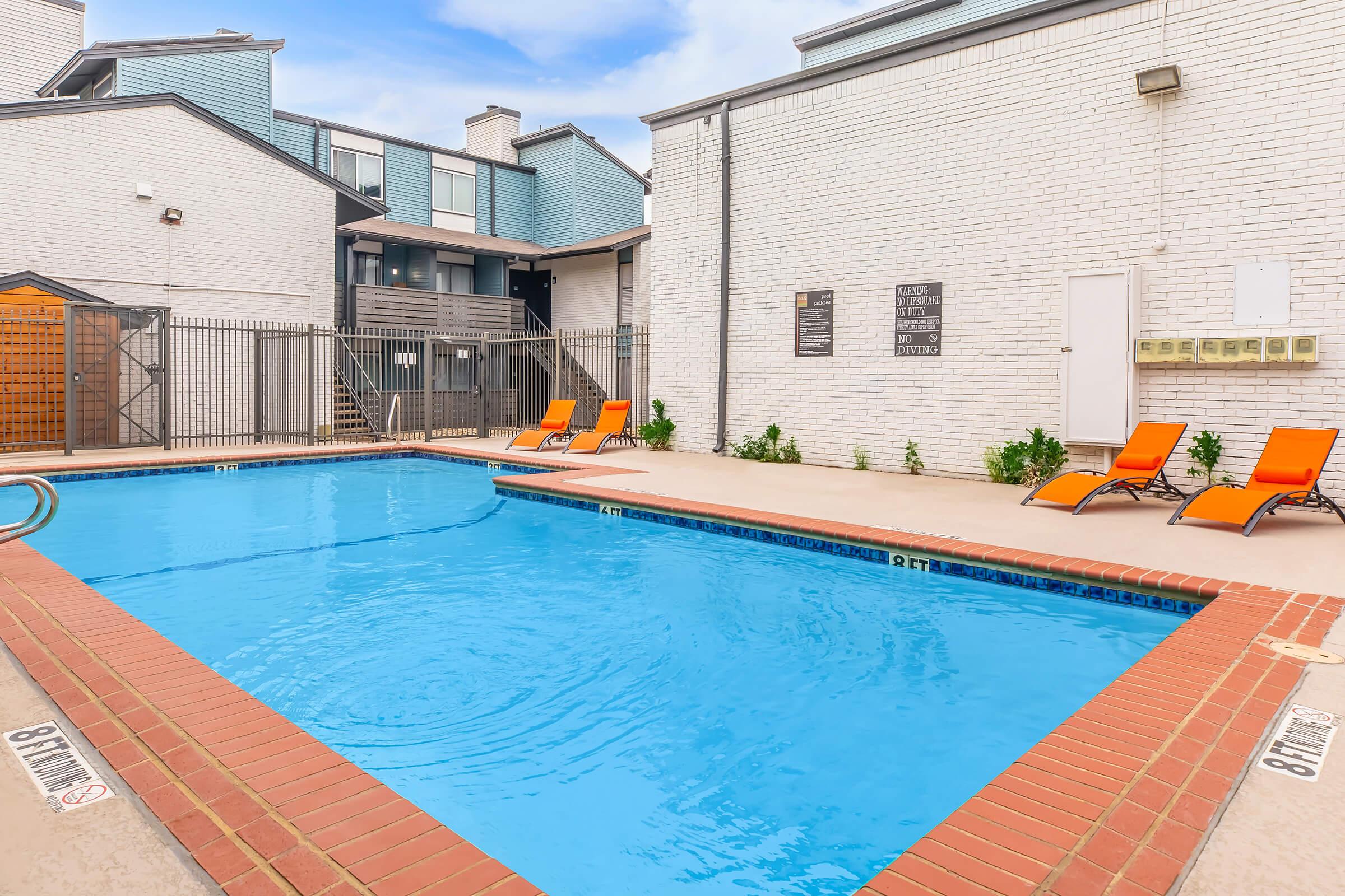 A clear blue swimming pool surrounded by a tan deck and orange lounge chairs. The pool area is enclosed by a fence, with a few buildings in the background. The sky is partially cloudy, and the setting gives a relaxed, inviting atmosphere.