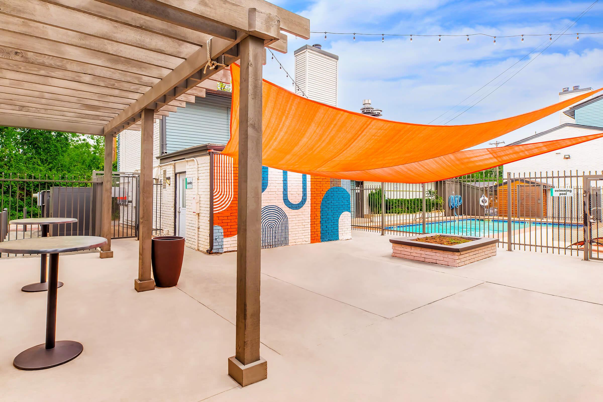 Outdoor pool area featuring a modern design with a vibrant orange shade sail overhead. There are round tables with chairs, a colorful mural on the wall, and a pool behind a gated fence. The space is bright and inviting, surrounded by greenery and a clear blue sky.