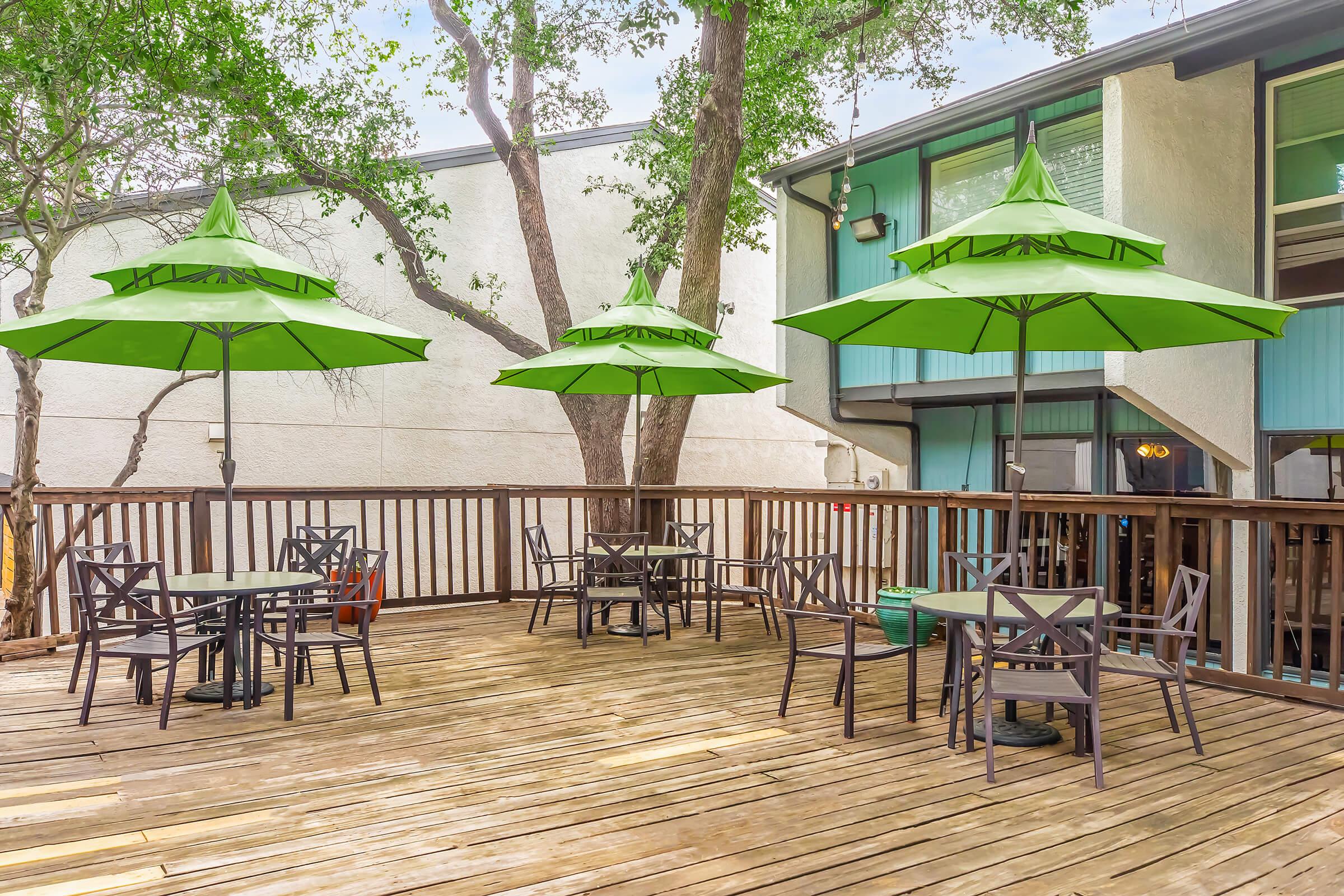 A wooden deck with several black tables and chairs, each covered by bright green umbrellas. Surrounding trees provide shade, and the background features a building with large windows. The scene is tranquil and inviting, perfect for outdoor dining or relaxation.