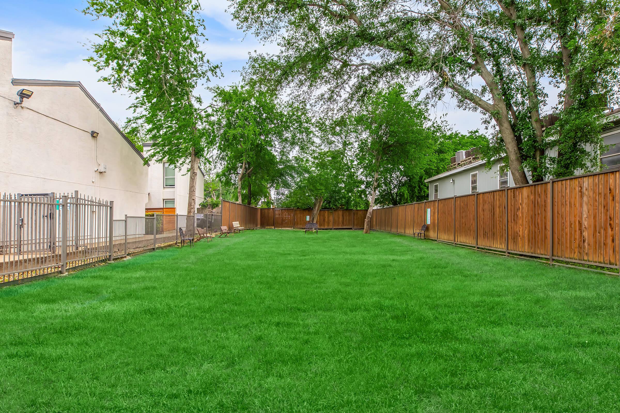 A spacious green lawn is bordered by wooden fencing and trees, with a few benches visible in the background. The scene is peaceful, suggesting a well-maintained outdoor area suitable for relaxation or play. Houses can be seen on the right side.