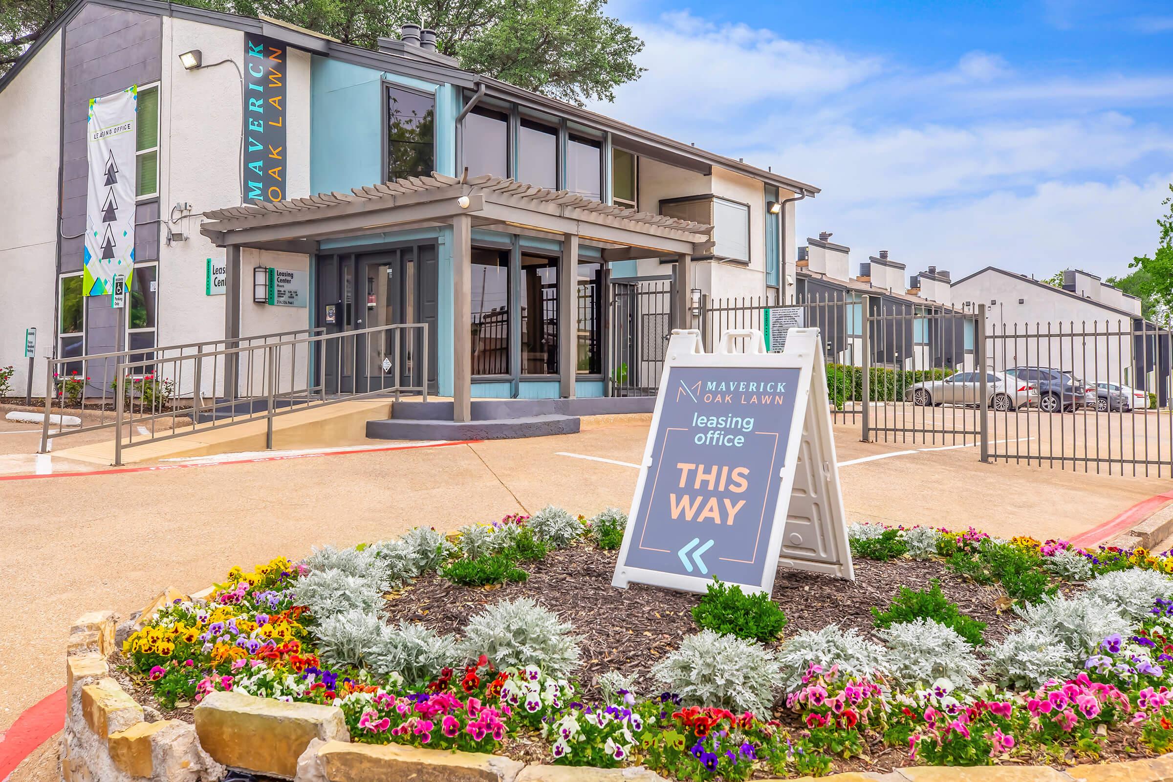 Leasing office exterior at Maverick Oak Lawn, featuring a directional sign that says "THIS WAY." The entrance has a landscaped area with colorful flowers and greenery, along with a modern building in the background.