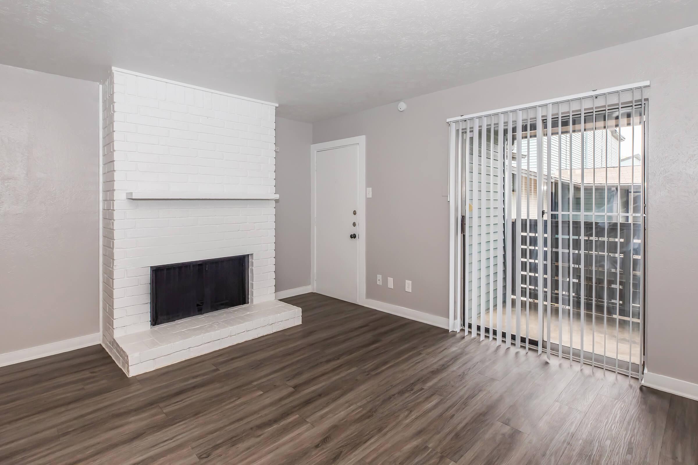 A modern living room featuring a white brick fireplace, light gray walls, and wooden laminate flooring. There is a sliding glass door with vertical blinds leading to an outdoor area, and a white door on the left side of the image. The space is bright and inviting with a minimalistic design.