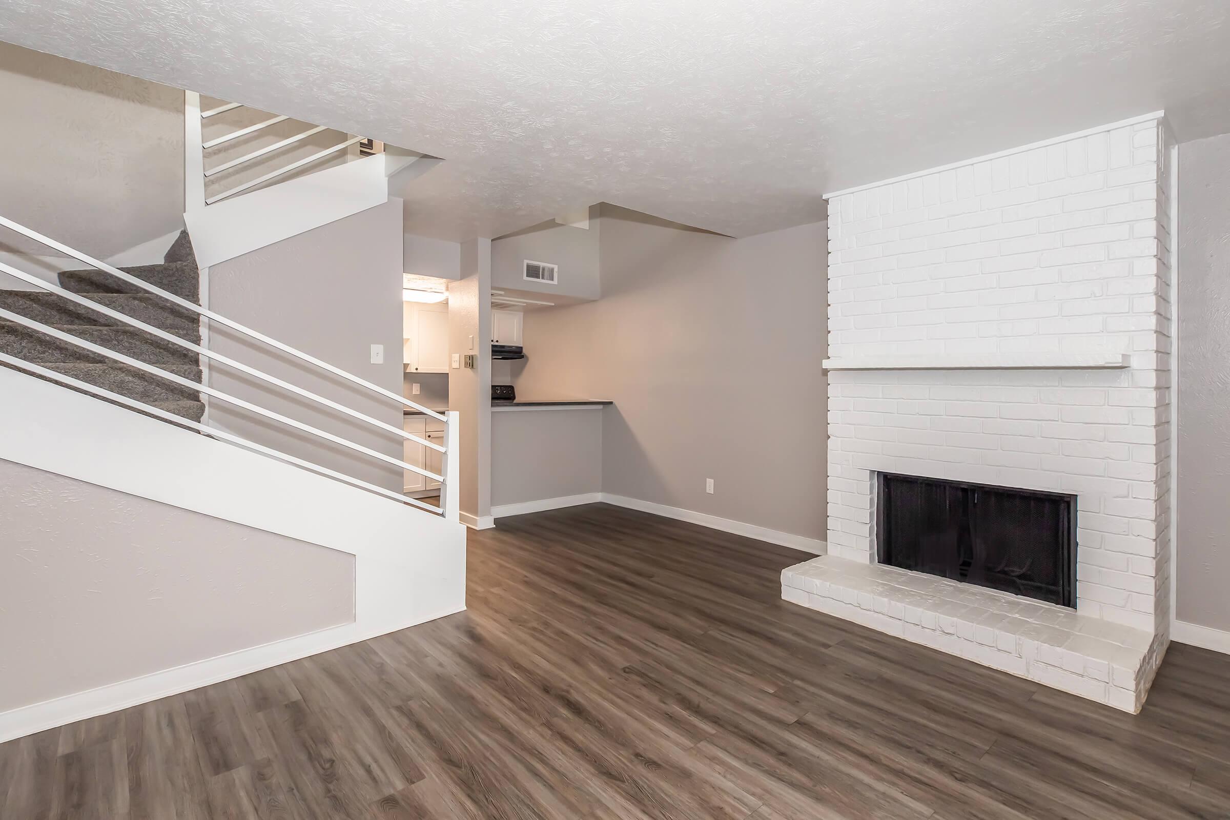 A modern living space featuring a white brick fireplace, laminate flooring, and neutral walls. A staircase is visible on the left, leading to an upper level. The open layout connects to a kitchen area in the background, creating a spacious and inviting atmosphere.