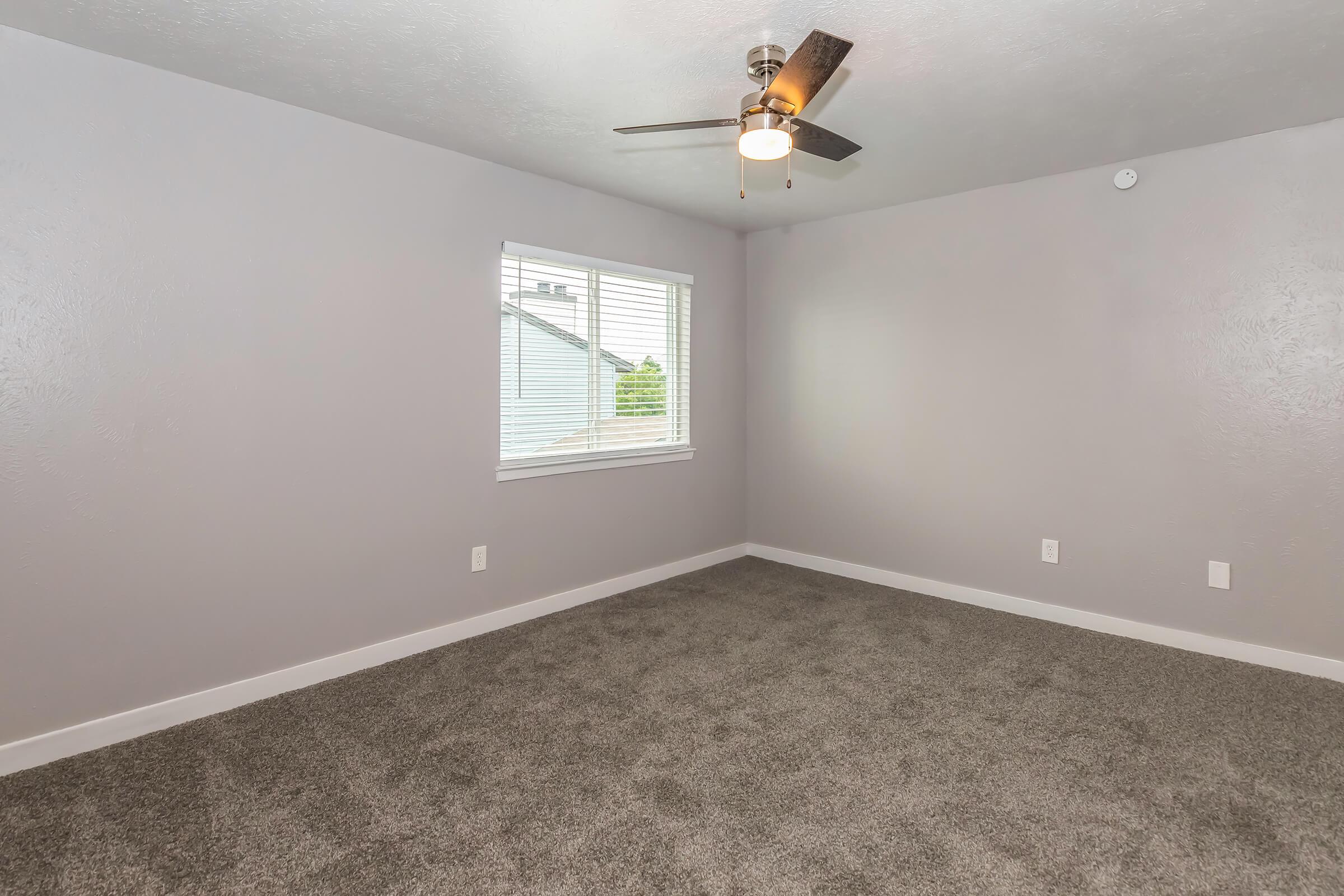 A spacious, light gray bedroom featuring a ceiling fan and a window with blinds. The floor is covered with plush, light-colored carpet, and the walls are painted in a soft gray hue. A small corner shows a glimpse of the outside through the window, creating a bright and inviting atmosphere.