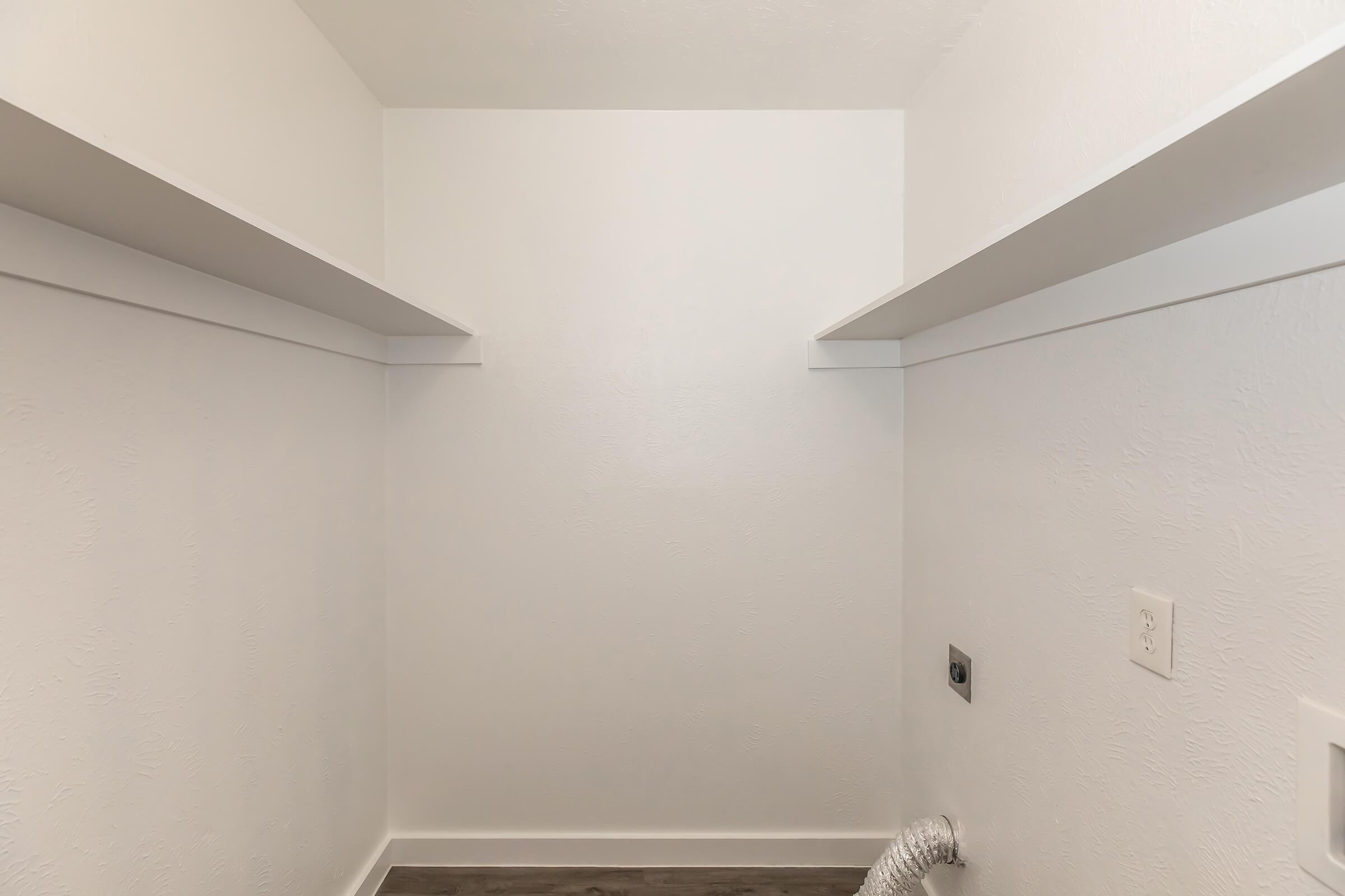 A clean, empty closet with white walls and two shelves. The floor is a light wood laminate, and there is a vent or duct on the right side. The space appears uncluttered and ready for organization or storage.
