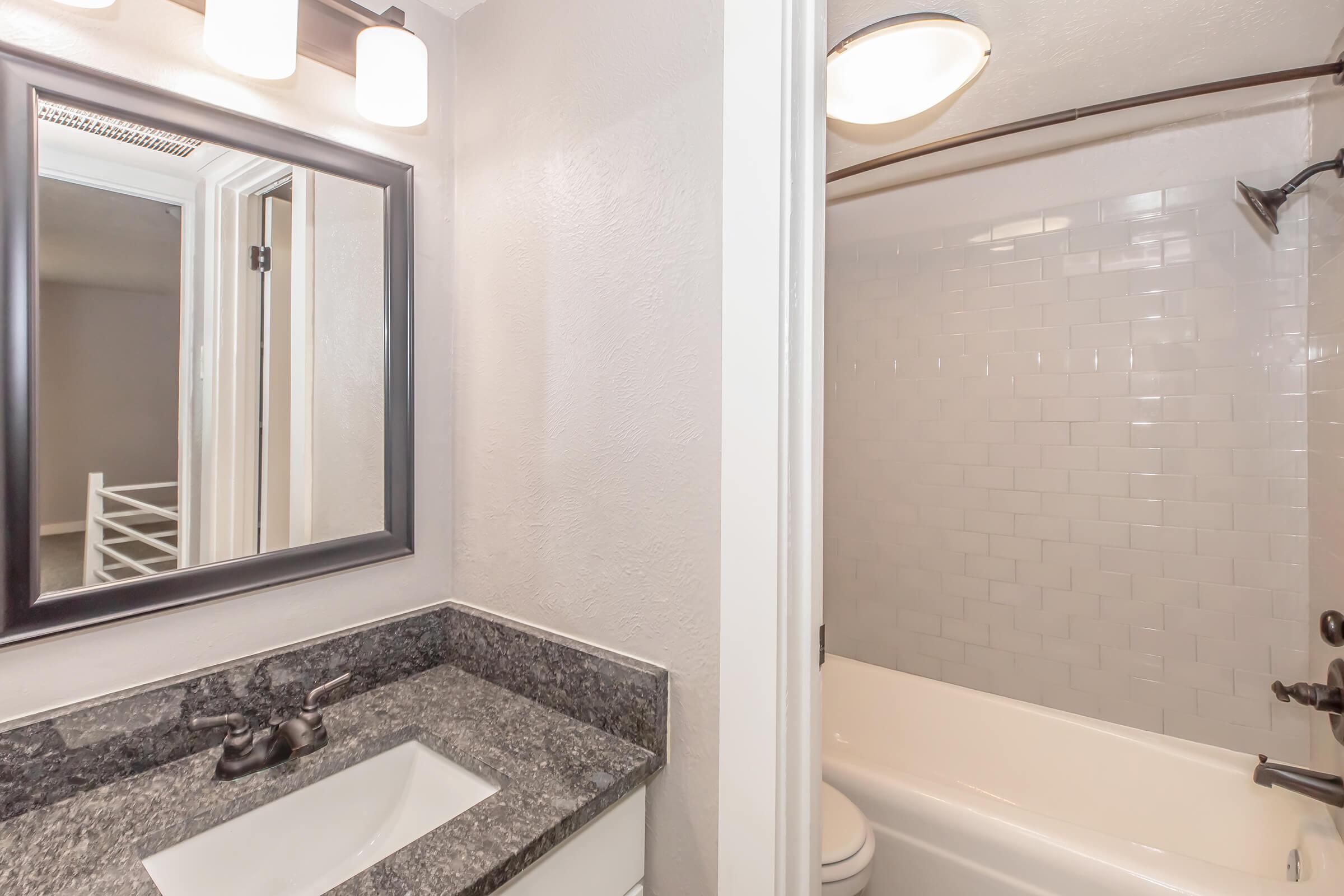 Modern bathroom featuring a dark granite countertop with a white sink, a large mirror with a black frame, and overhead lighting. To the right, a shower/tub combination with white subway tiles. The space appears bright and clean, with neutral-colored walls and a glimpse of another room in the background.
