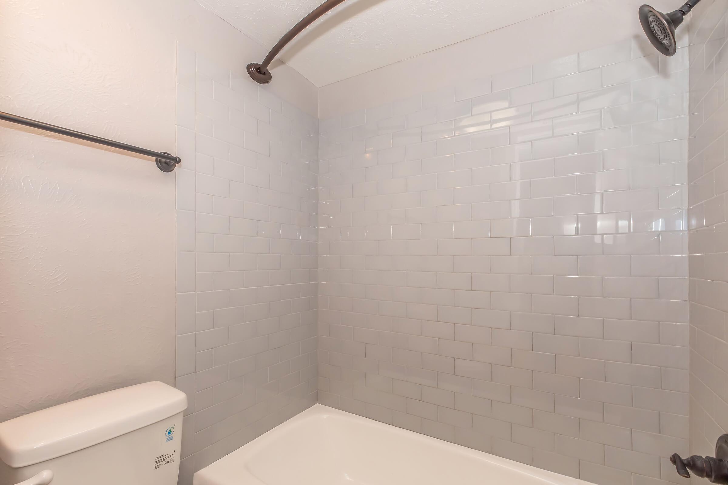 A clean, modern bathroom featuring a shower area with gray subway tiles, a white bathtub, and a white toilet. The space has a simple design with a curved shower rod and a showerhead, lending it a fresh and contemporary feel.