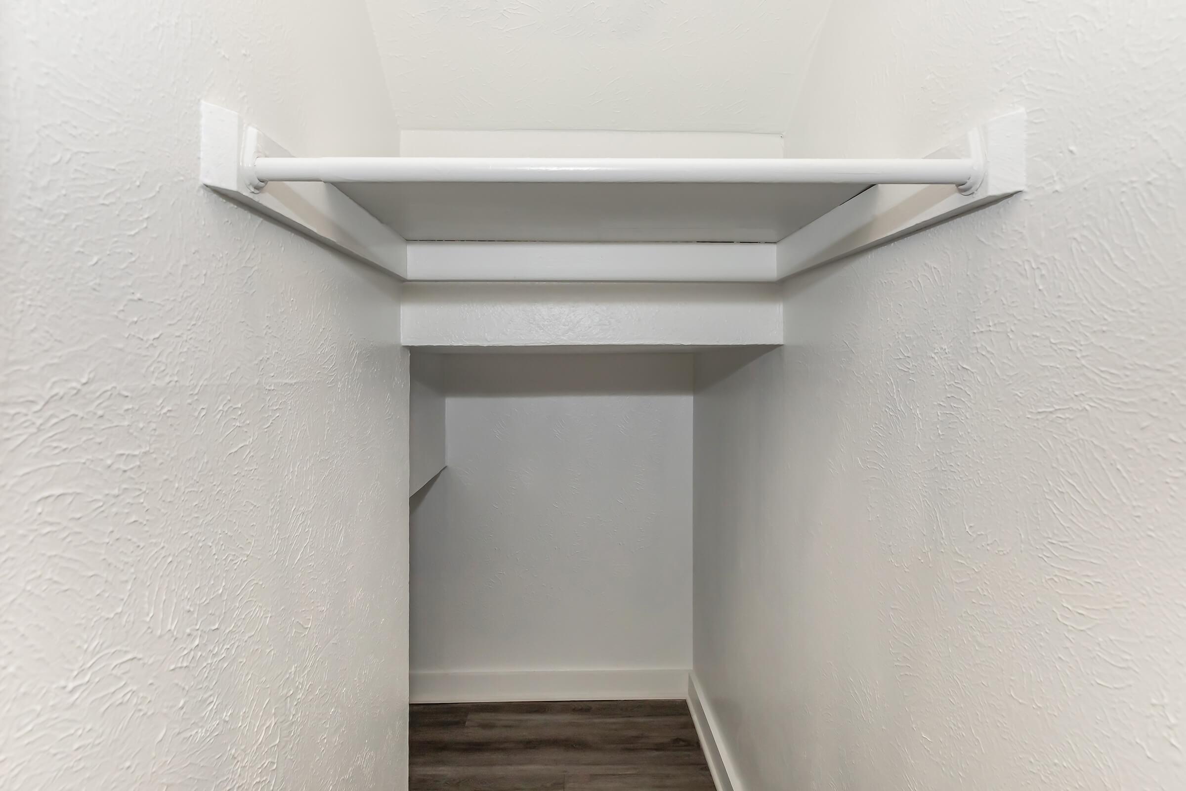 A view of a small, empty closet with white walls and a hanging rod. The floor is covered with dark wood laminate, and there is a shelf above the rod. The design is simple and minimalistic, providing a clean space for storage.