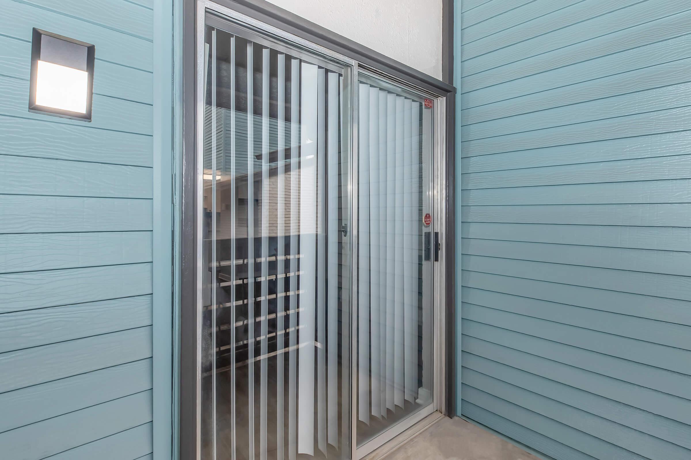 Sliding glass doors with vertical blinds leading to a patio area, set against a light blue wooden wall. A small wall-mounted light fixture is visible on the left side. The area appears modern and clean, providing a view into a storage space beyond the doors.
