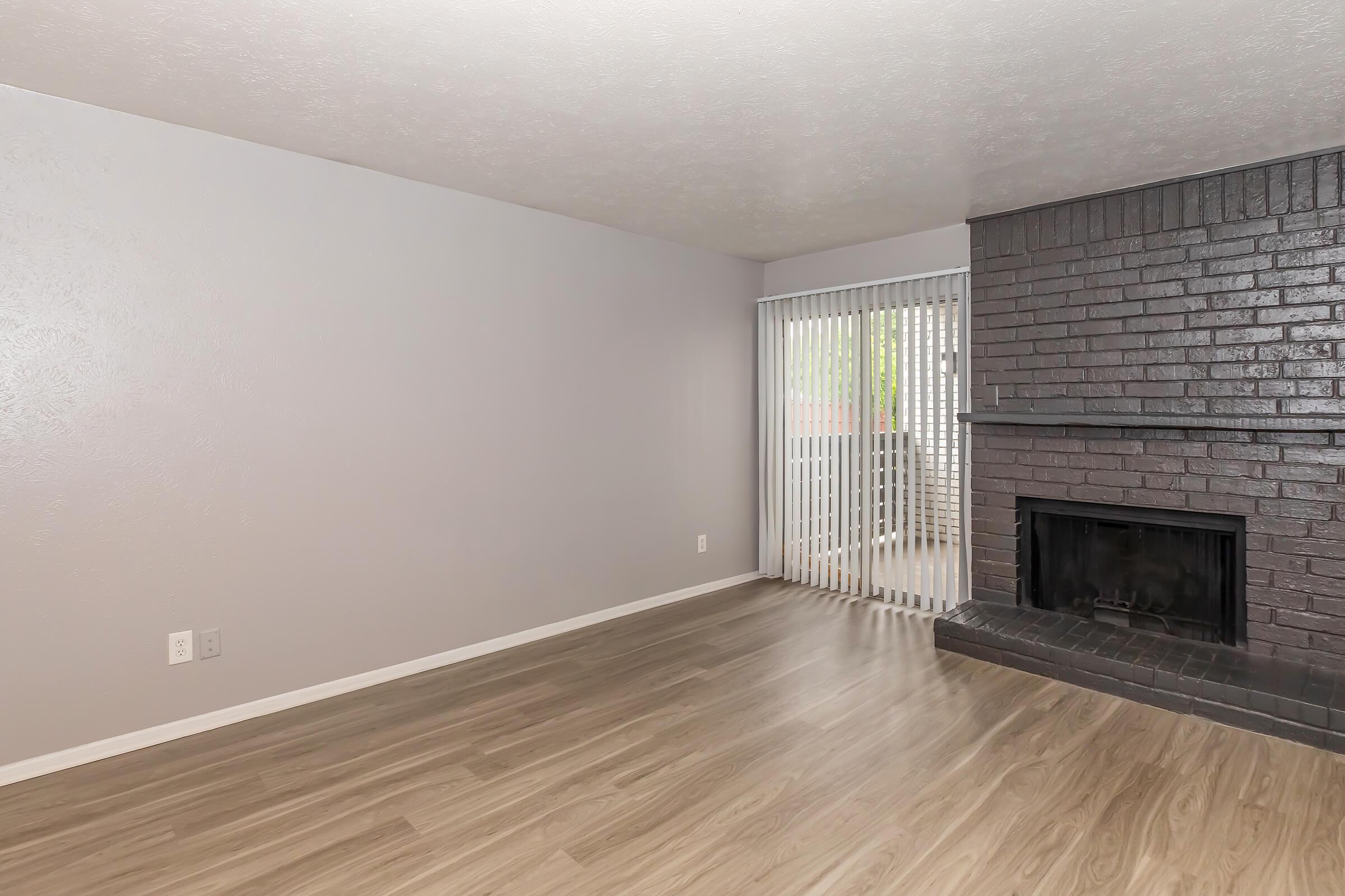 A spacious, empty living room featuring light-colored walls, a brick fireplace, and large windows with vertical blinds. The floor is covered in light wooden laminate. The overall atmosphere is bright and airy, perfect for personalization and decor.