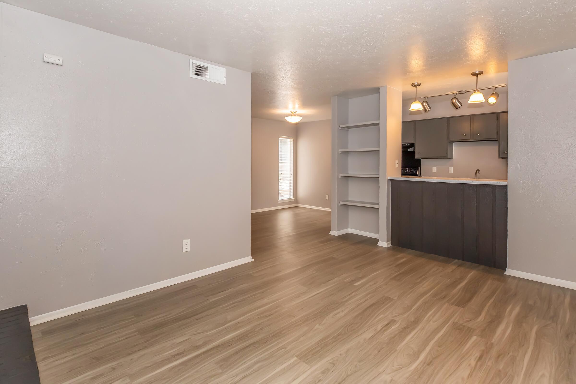 Interior view of a modern, spacious living area with light-colored laminate flooring. The room features grey walls, a large window allowing natural light, and an open layout connecting to a kitchen with three pendant lights and dark cabinetry, along with shelves for storage.