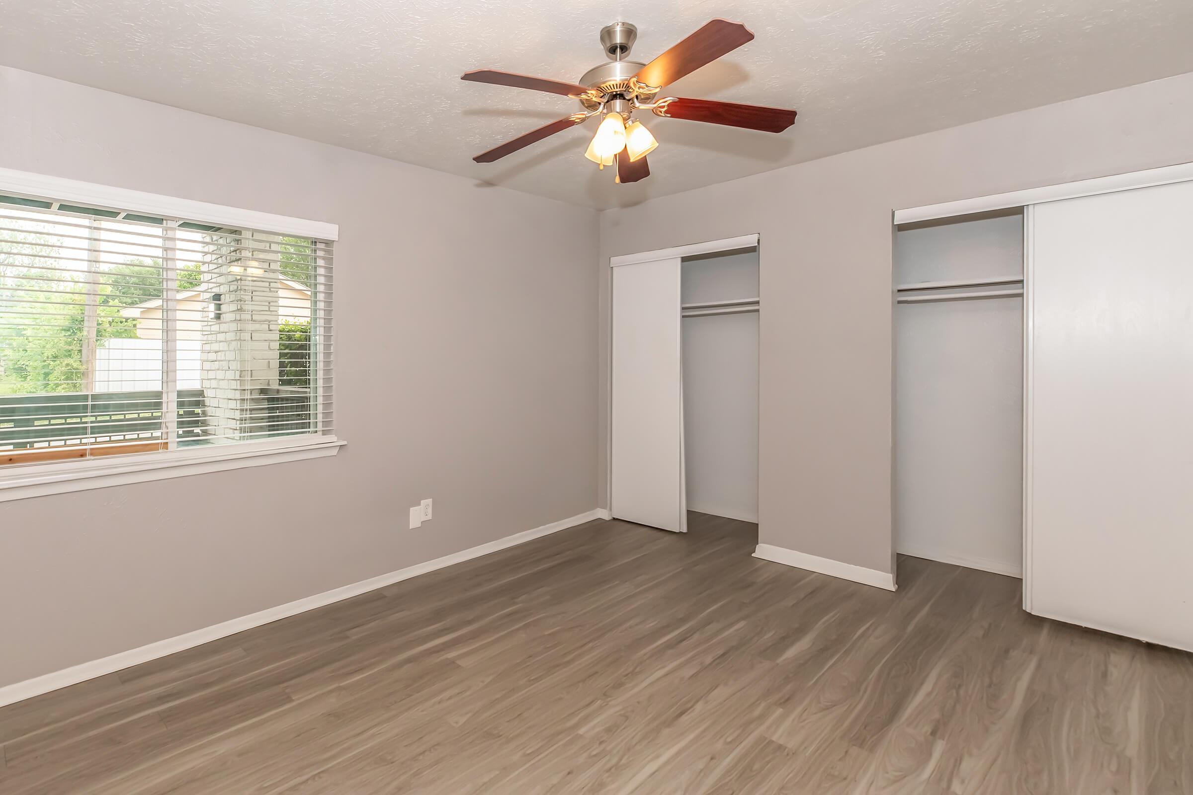 A light gray bedroom featuring a ceiling fan with three lights. There are large windows with white blinds allowing natural light. Two closets with sliding doors are visible on one side, and the floor is covered with light wood laminate. The walls are smooth and neutral in color.