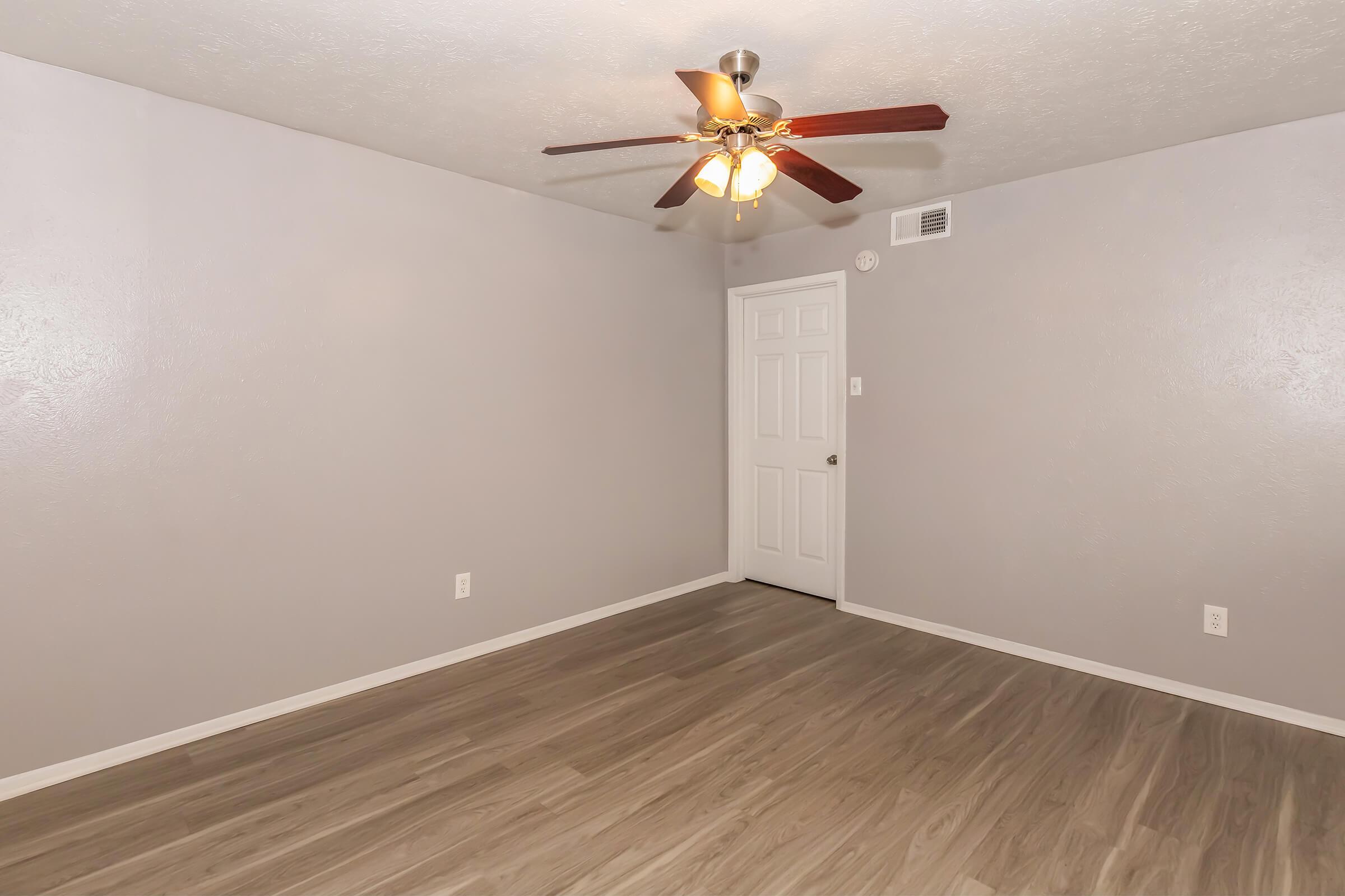 A sparsely furnished room with light gray walls and a ceiling fan with four light bulbs. The floor is made of light-colored laminate. A white door is visible on the right side of the image, leading to another room or hallway. The space appears clean and well-lit, suitable for various purposes.