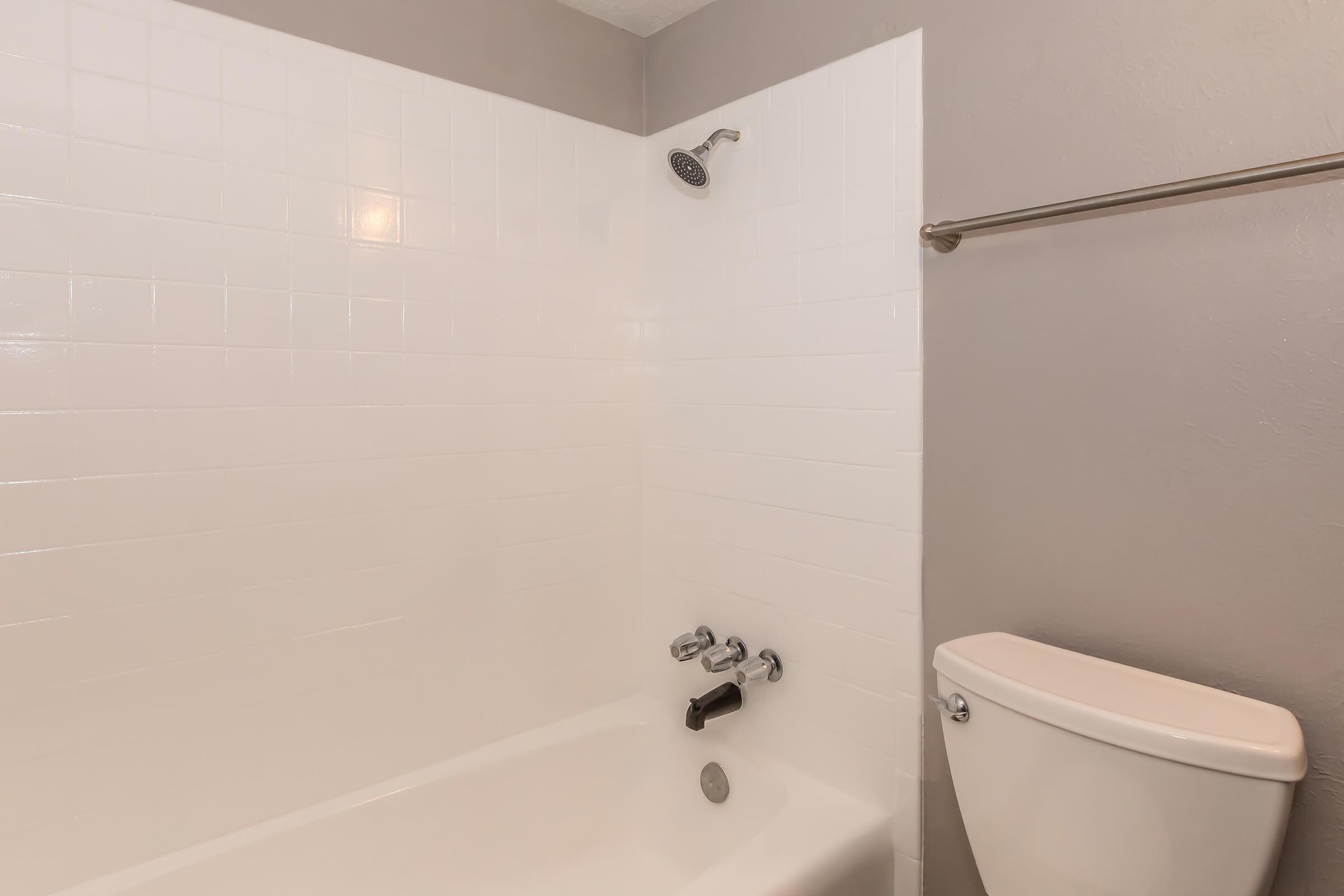 A clean, modern bathroom featuring a white bathtub with a silver faucet and showerhead. The walls are tiled in white, and there is a simple toilet adjacent. The overall design is minimalistic, with a neutral color scheme and no decorative elements.