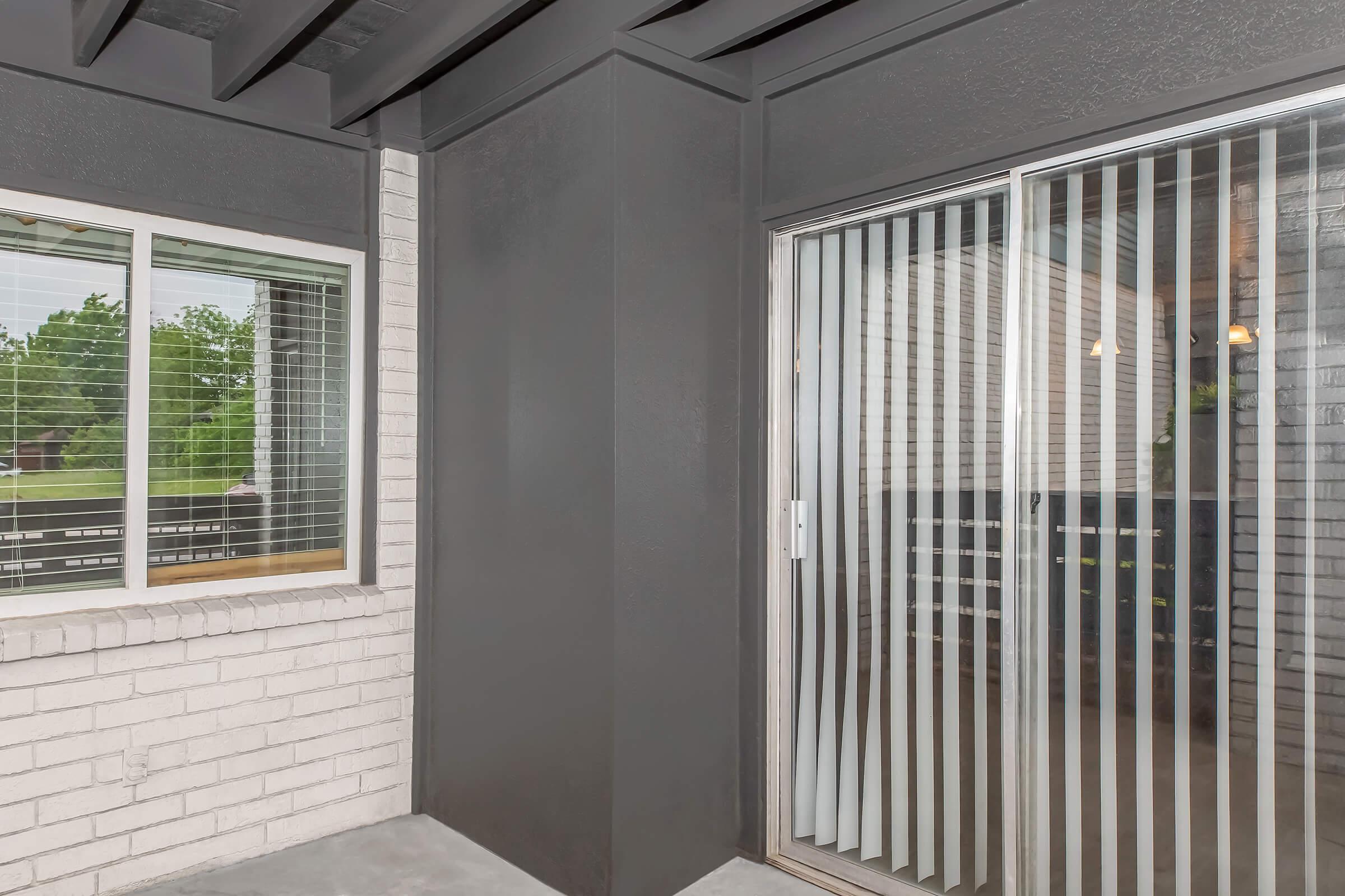 Interior view of a modern balcony with a sliding glass door featuring vertical blinds. The walls are painted in a dark color, contrasting with a light-colored brick wall. A window with blinds is visible on the left side, allowing natural light to brighten the space. Greenery can be seen outside.