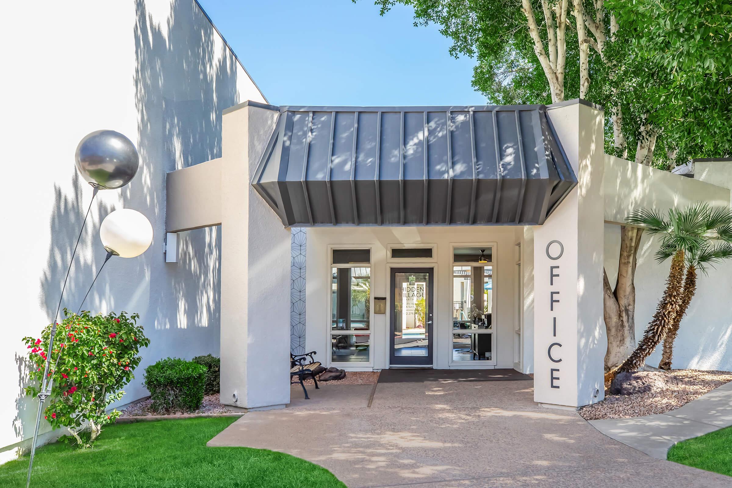 Entrance to an office building with a modern design. The façade features a gray metal roof and large glass doors. A sign reads "OFFICE" on the side. Lush greenery, including a small palm tree and colorful flowers, is visible. The walkway is paved, leading to the entrance. Two decorative balloons are placed nearby.