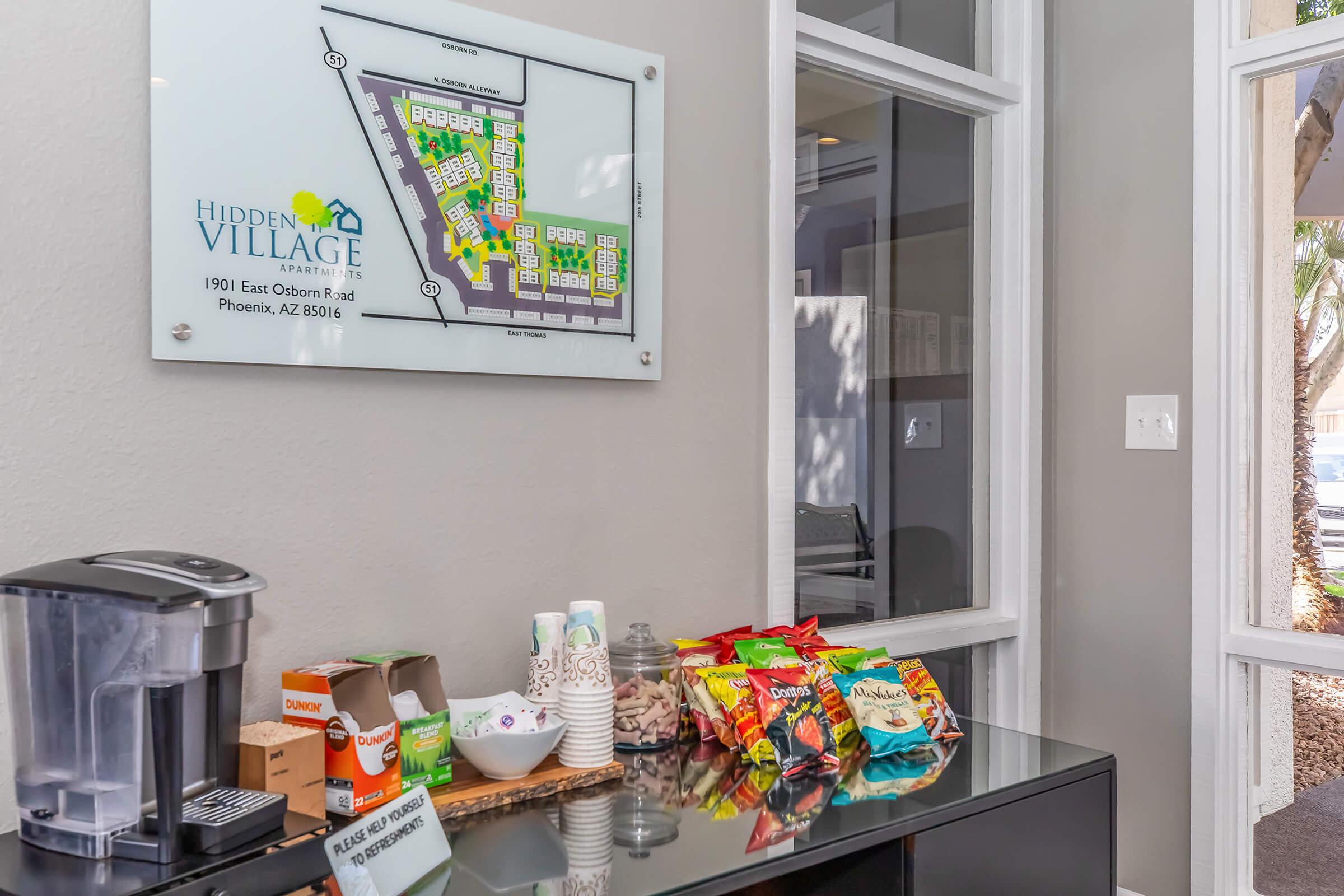 A lobby area featuring a map of Hidden Village Apartments on the wall, with a coffee station and snack display. The table has various snacks, coffee supplies, and a sign indicating amenities. Natural light comes from nearby windows, creating a welcoming atmosphere.