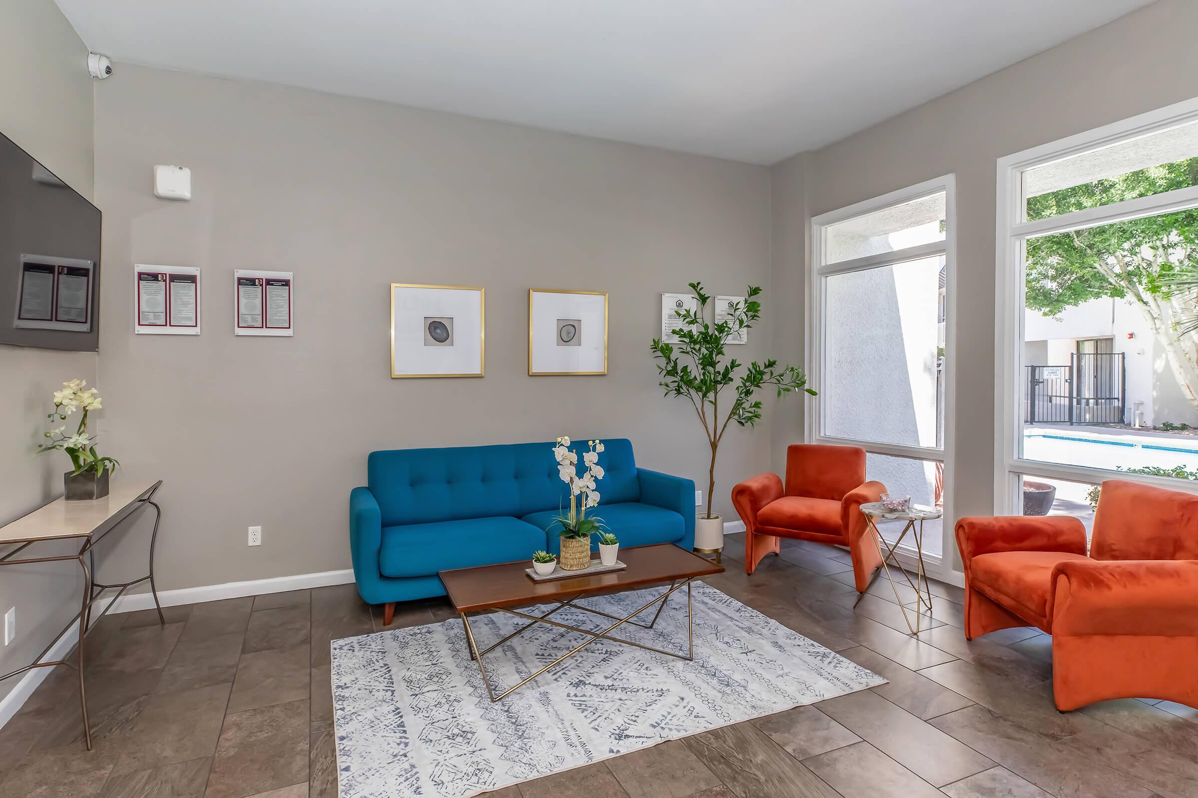 A modern living room featuring a stylish blue sofa and two orange armchairs, complemented by a coffee table and a decorative rug. Large windows allow natural light to fill the space, with greenery visible outside, creating a bright and inviting atmosphere.