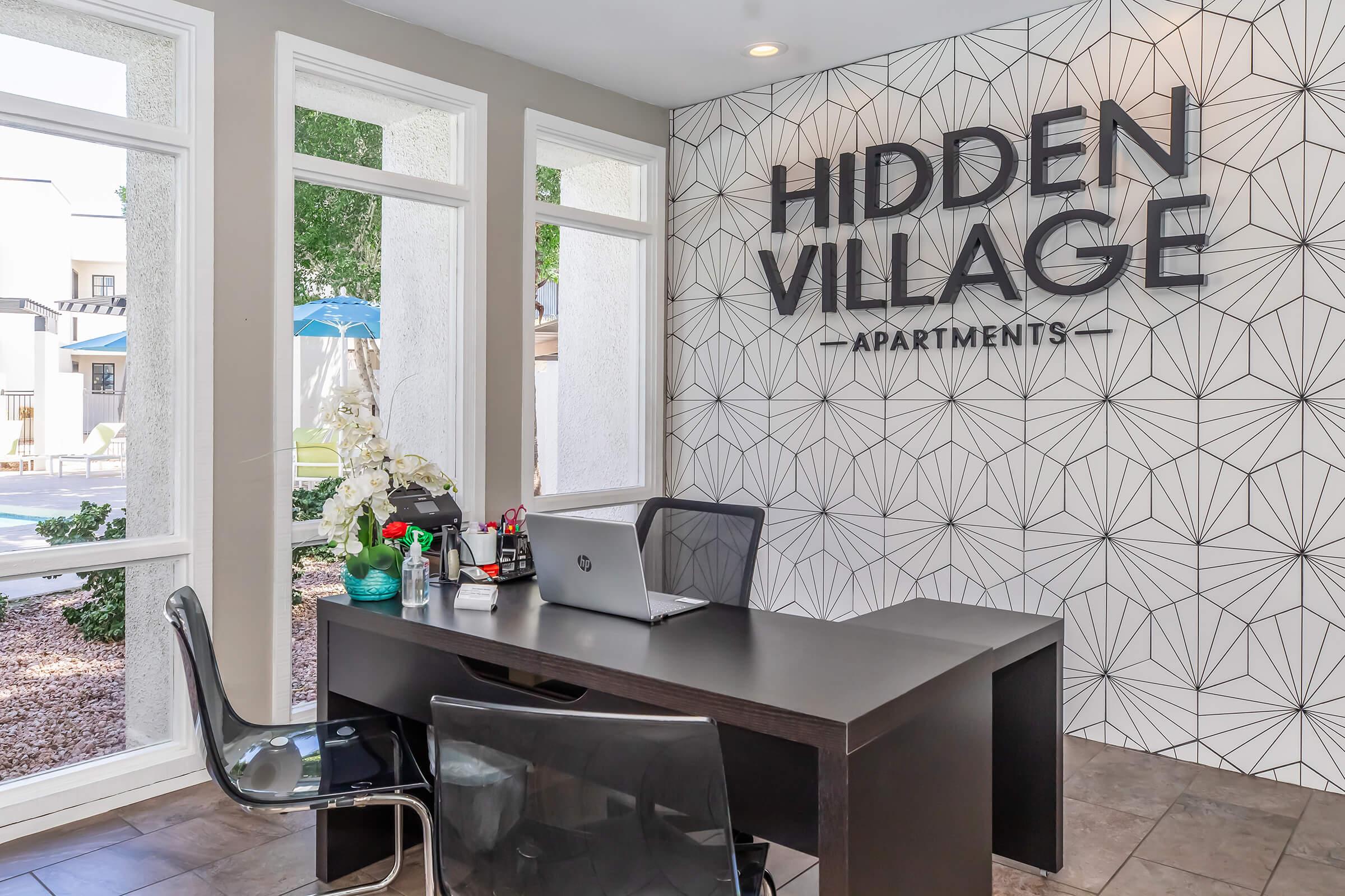 Interior view of a stylish apartment leasing office featuring a modern black desk with a laptop, a chair, and decorative items. The wall behind displays the text "Hidden Village Apartments" with a geometric design. Large windows allow natural light to illuminate the space.