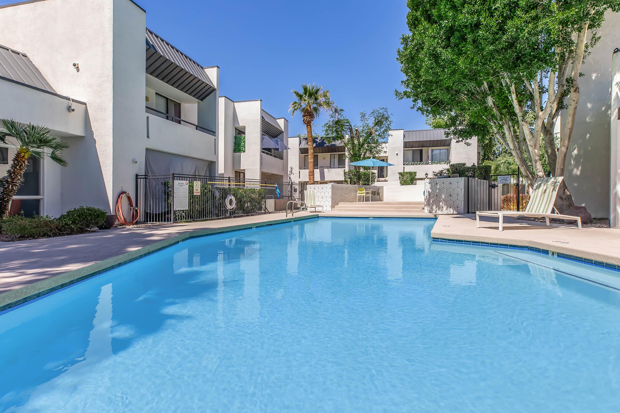 A clear blue swimming pool surrounded by modern apartment buildings. Lush green trees and palm plants adorn the area. A few lounge chairs and a table with an umbrella provide seating by the pool. The sky is bright and sunny, creating a tranquil and inviting outdoor atmosphere.