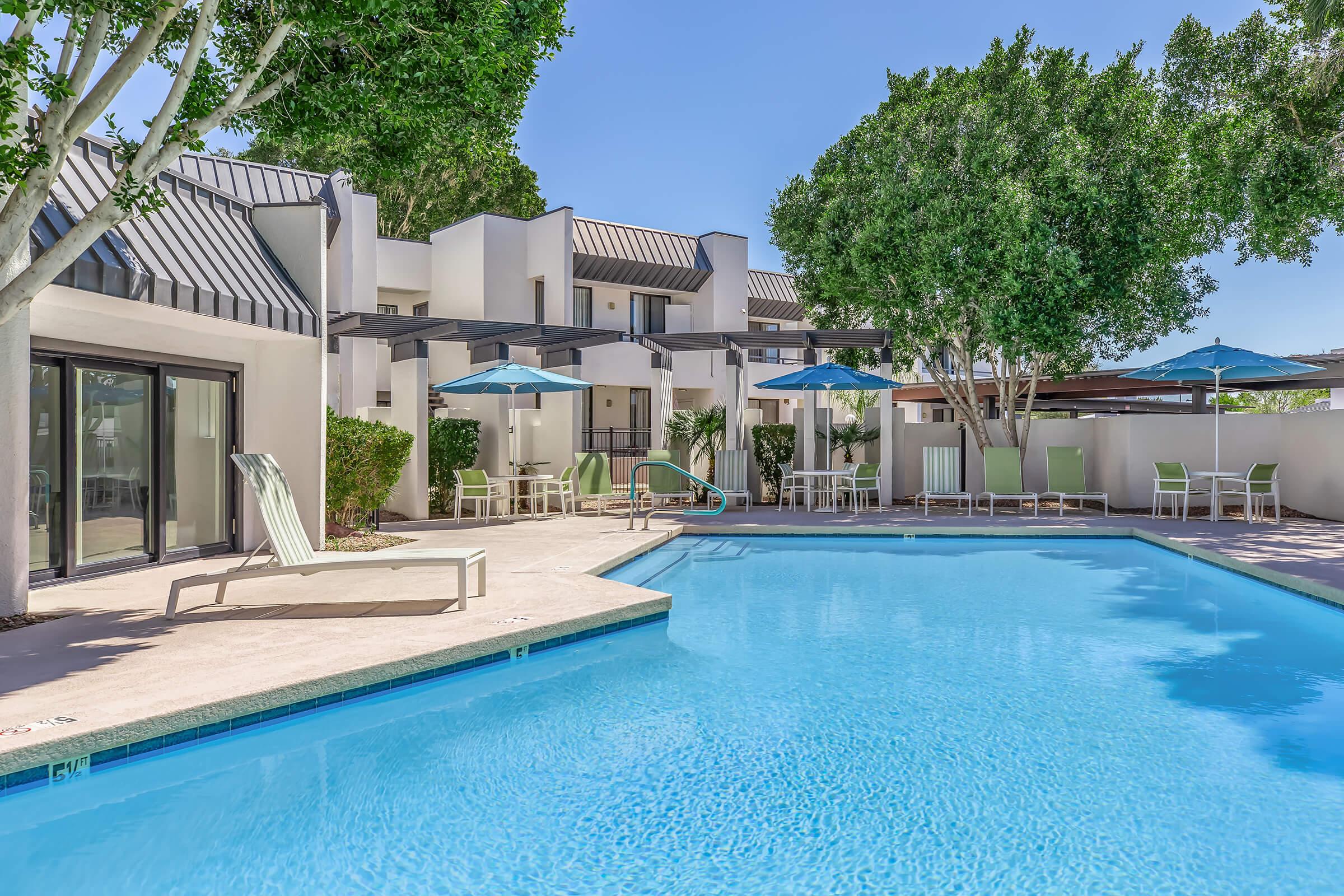 A bright swimming pool surrounded by lounge chairs and blue umbrellas, with modern buildings and lush greenery in the background. The scene conveys a relaxing atmosphere, ideal for leisure and entertainment.
