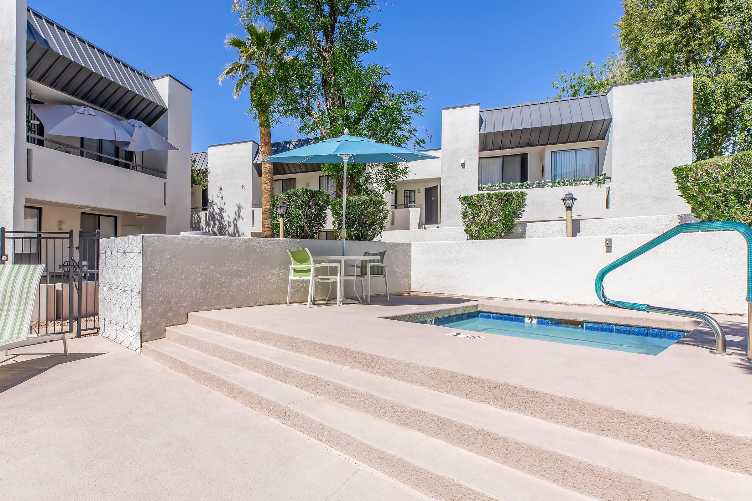 A sunny outdoor area featuring a small pool, surrounded by white walls and greenery. There's a table with a green chair under a blue umbrella and several white umbrellas nearby. The setting is part of an apartment complex, with clear blue skies above.