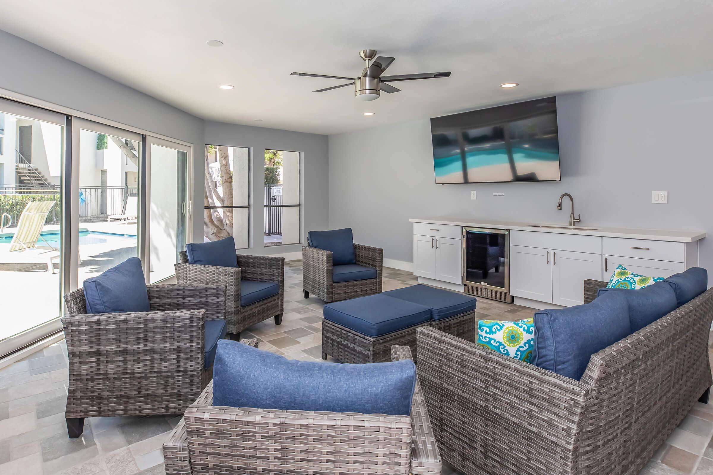 Cozy lounge area with four wicker chairs and a matching ottoman, all with blue cushions. A wall-mounted TV is above a modern cabinet with a mini refrigerator. Large glass sliding doors lead to a pool area, and the space features neutral walls and tiled flooring, creating a bright and inviting atmosphere.