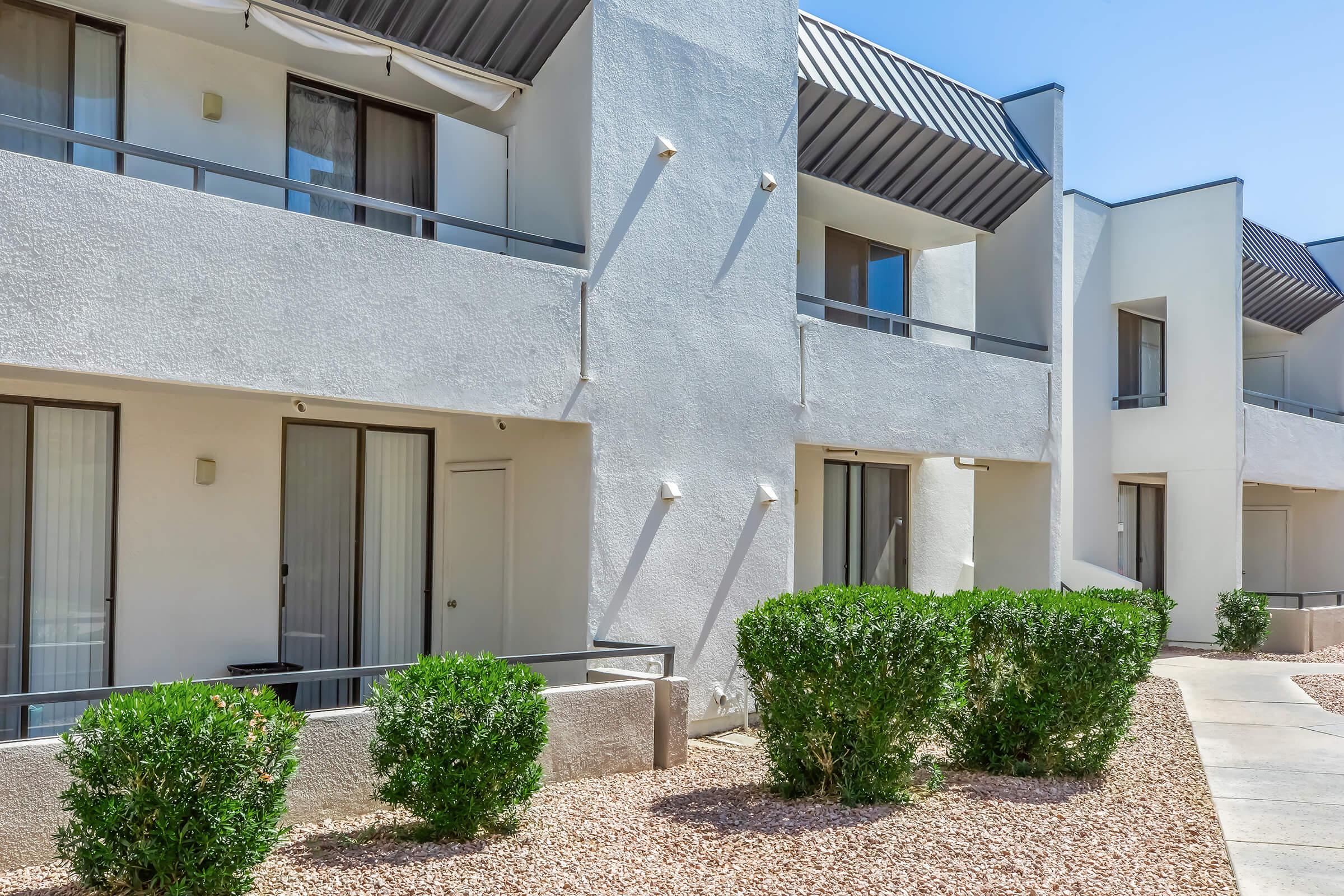 Modern building exterior featuring two-story white walls, large windows with sliding doors, and a walkway surrounded by low shrubbery and decorative gravel. The upper floors have balconies with shaded awnings, creating a clean and minimalistic architectural design under a clear blue sky.
