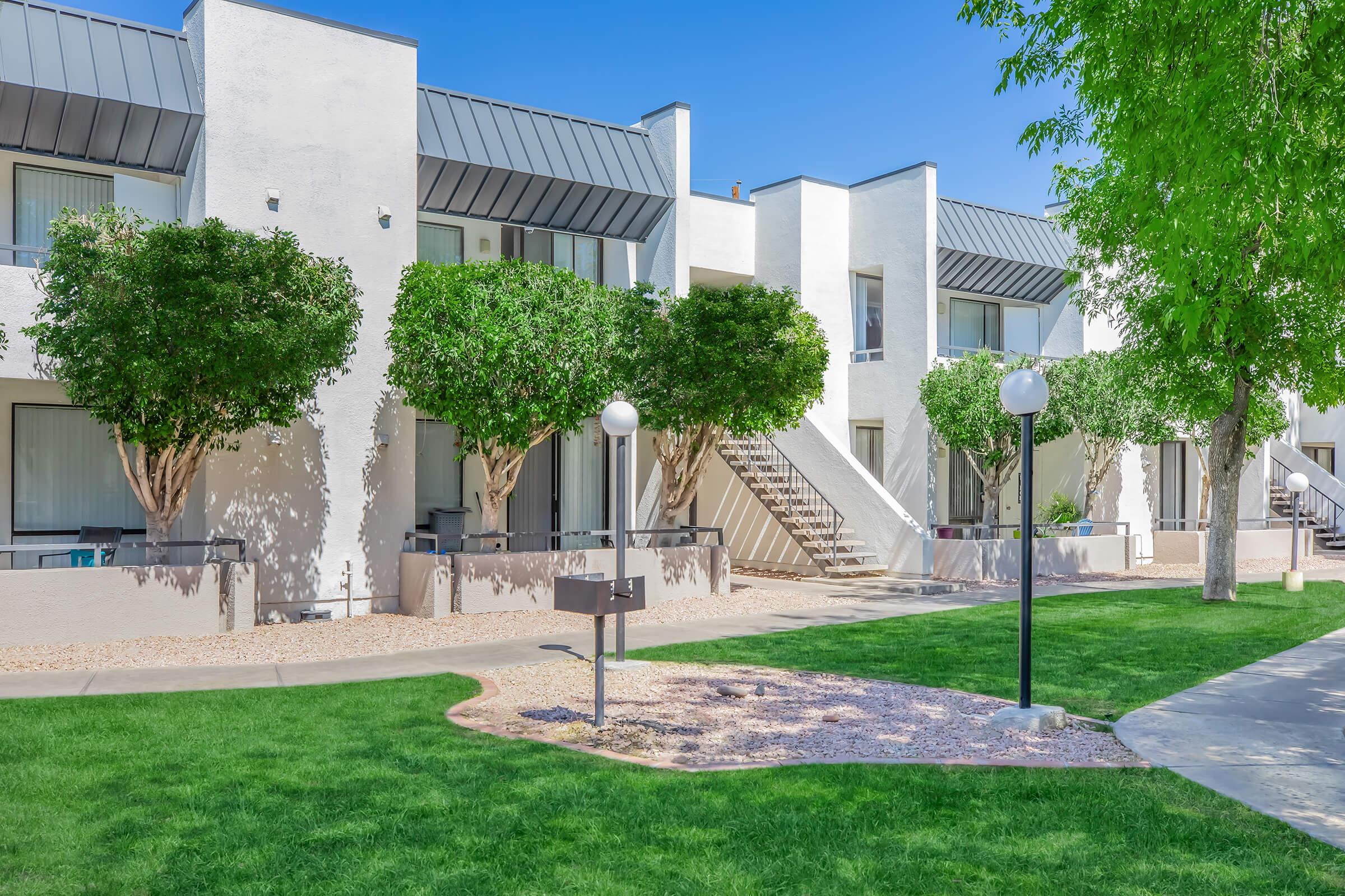 Two-story residential building with a modern design, featuring multiple units. Each unit has a small balcony and is surrounded by well-maintained trees and green lawns. The area is sunny, with clear blue skies, and includes decorative light posts along the pathway leading to the entrances.