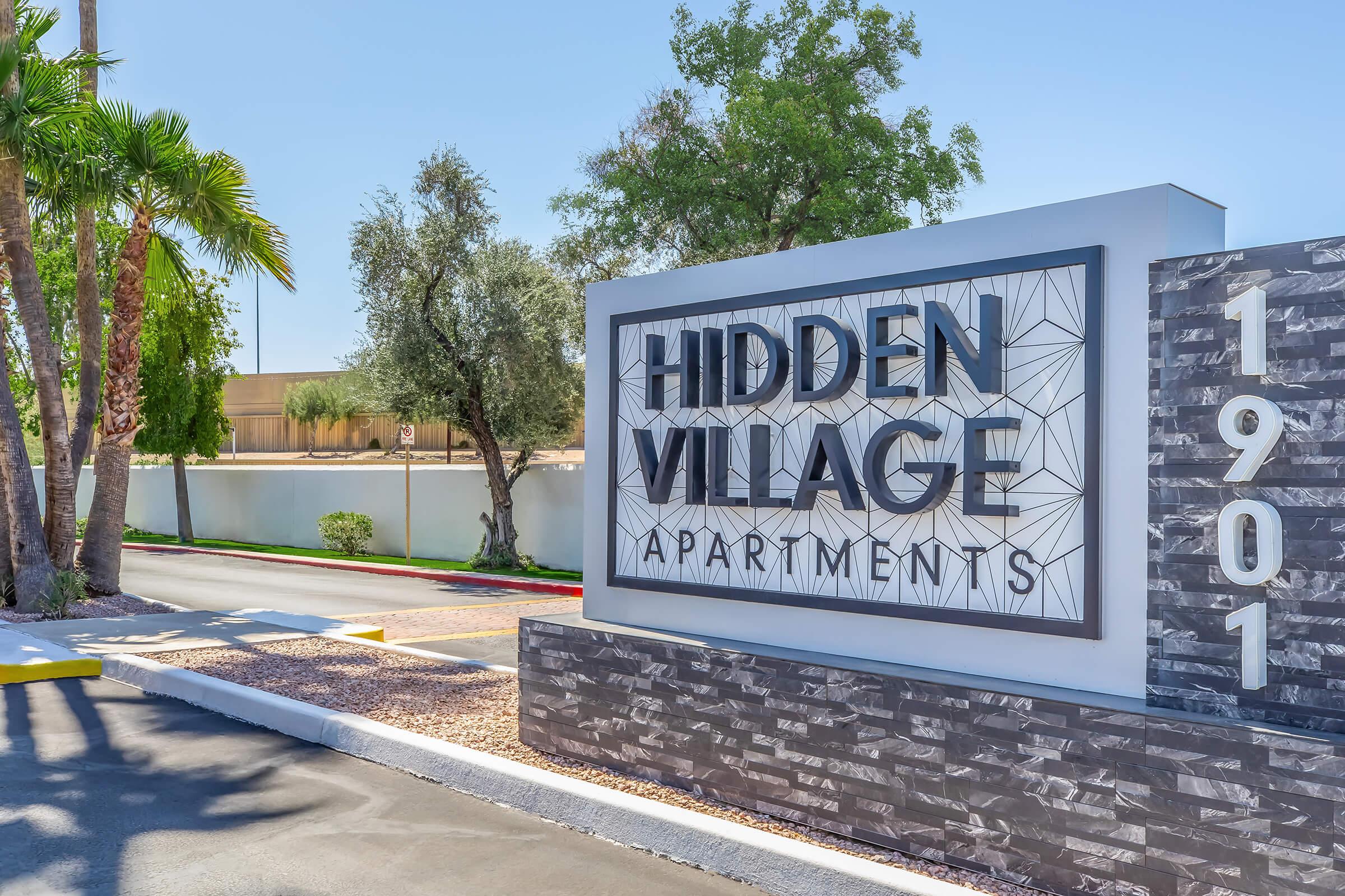 Sign for Hidden Village Apartments with modern design, featuring the name prominently displayed, surrounded by palm trees and landscaping. The background shows a clear blue sky and a driveway leading into the apartment complex.