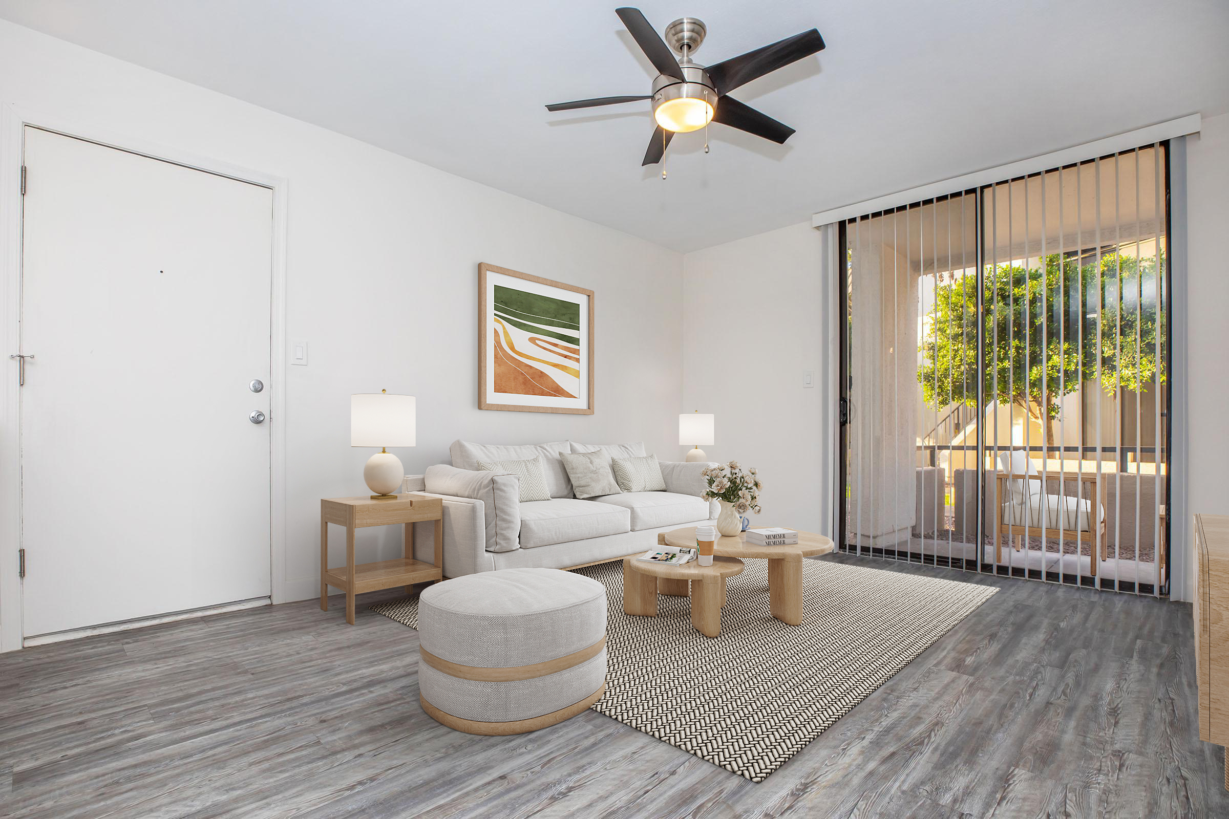 A light and airy living room featuring a white sofa with decorative pillows, a round coffee table with a vase of flowers, and a plush ottoman. The space has a textured area rug and is illuminated by two side lamps. A large sliding door opens to a patio with greenery visible outside.