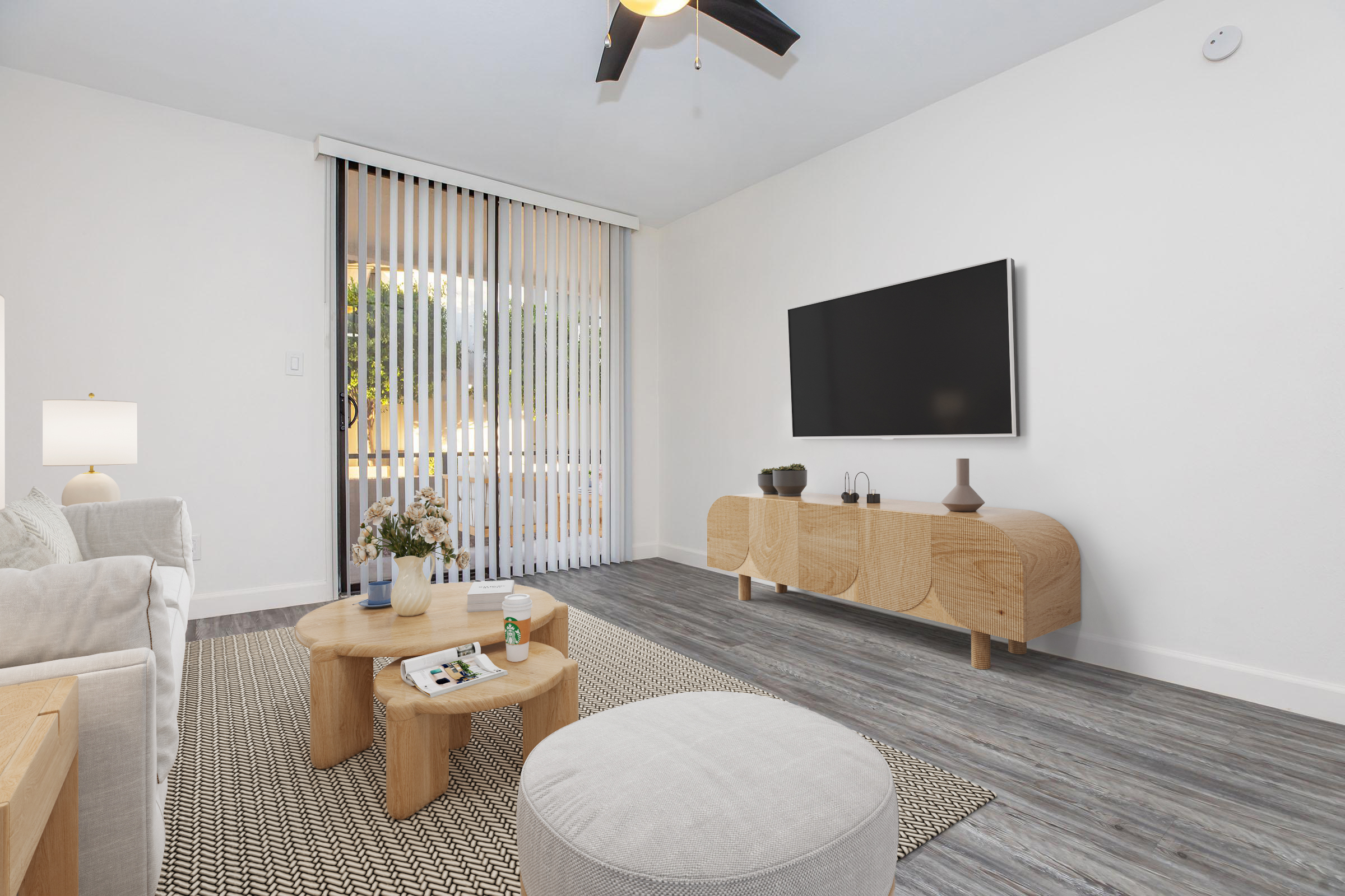 A bright, modern living room featuring a light-colored sofa, a round coffee table with decor, and a flat-screen TV mounted on the wall. A small lamp sits on a side table, and a large window with vertical blinds provides natural light. The flooring is a sleek wood-look design.